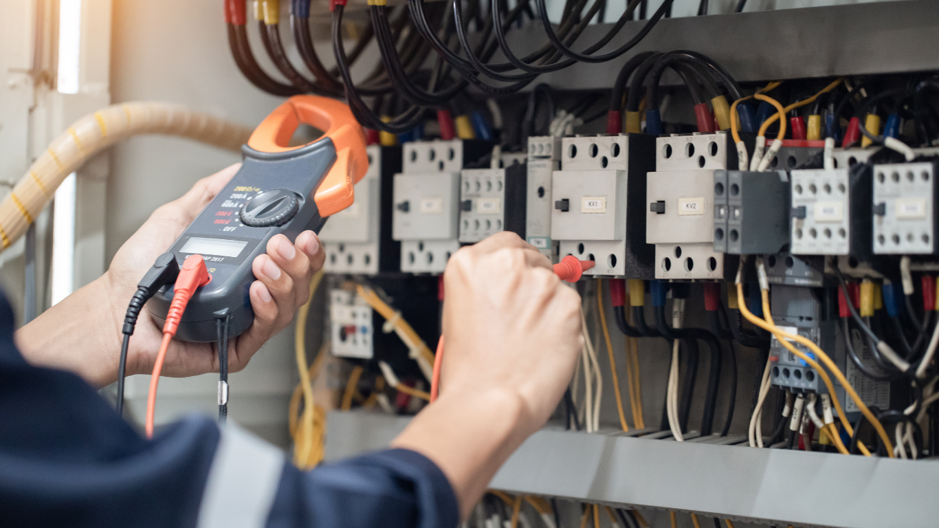 Technician testing electrical control panel with clamp meter and adjusting wiring.