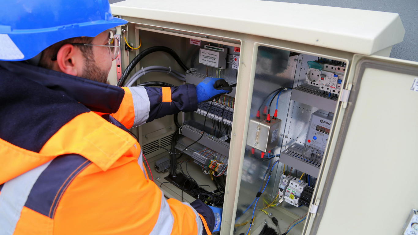 Technician in orange safety gear inspecting an open electrical control cabinet outdoors.