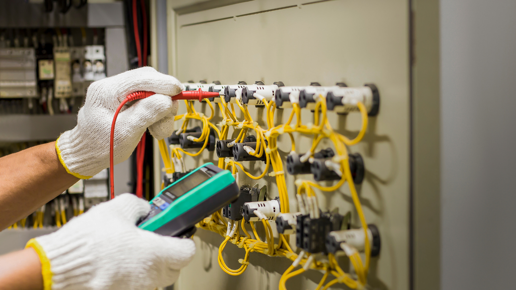 Gloved hands testing wiring in an electrical control panel with yellow cables and a multimeter