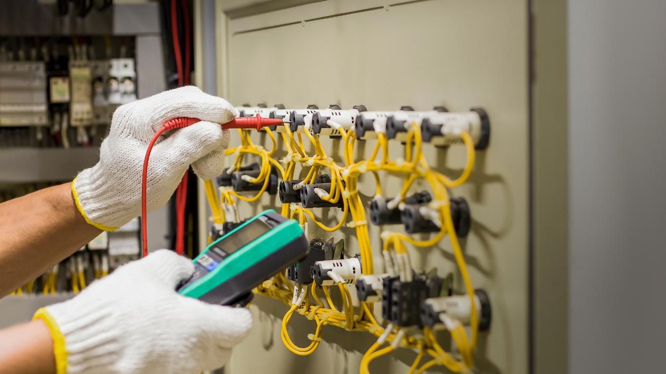 Gloved hands testing wiring in an electrical control panel with yellow cables and a multimeter