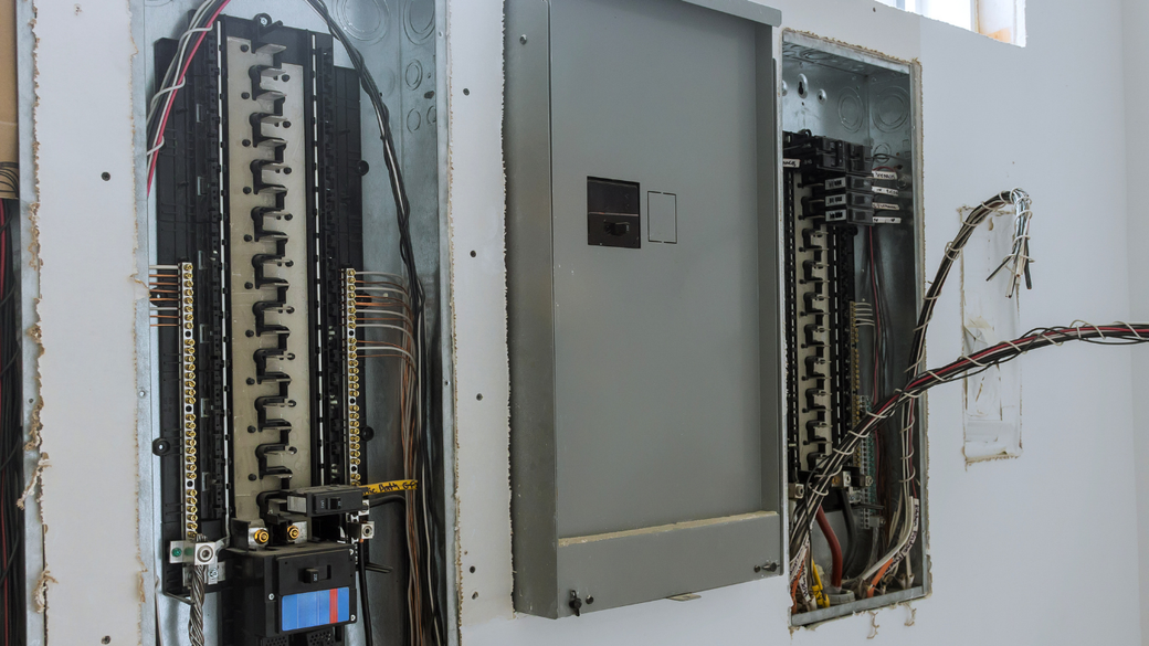 Open electrical breaker panels exposed in a wall, with wiring and a gray service box.