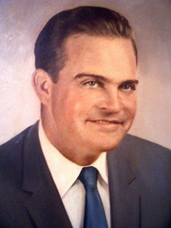 Studio portrait of a smiling man in a dark suit, white shirt, and blue tie, against a soft pink background