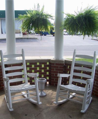 Two white rocking chairs on a porch with hanging fern baskets and columns in the background.