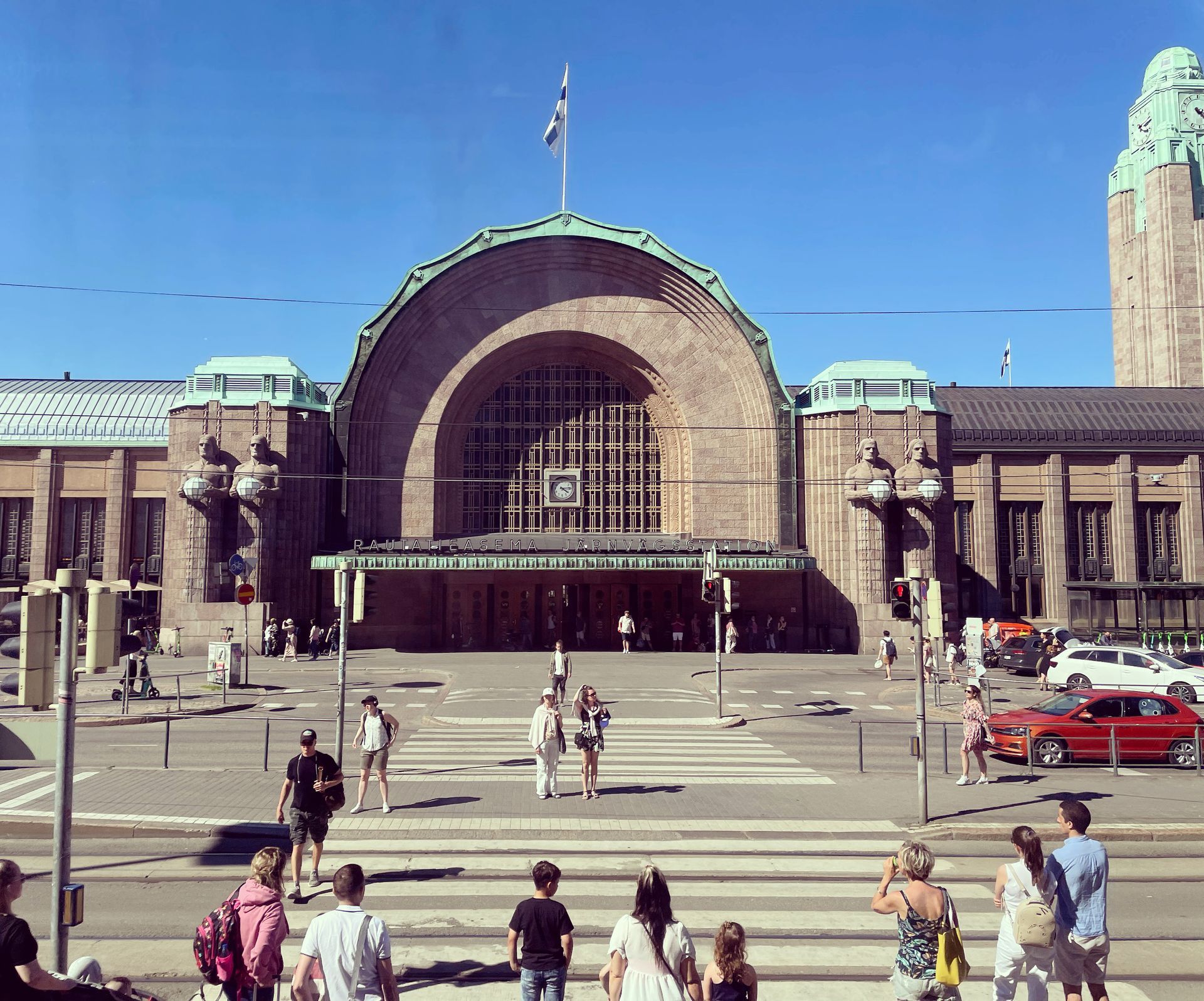 Helsinki Central Station exterior on a sunny day with people crossing a street. Finnish flag flies above.