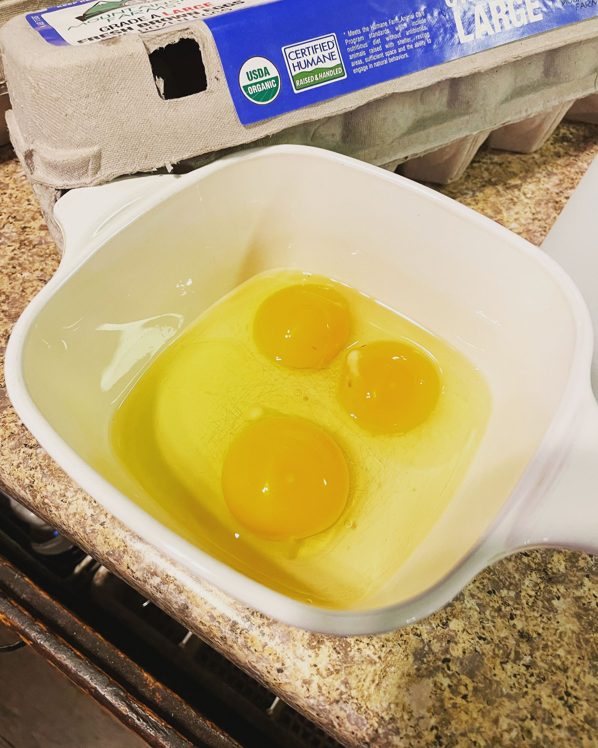 Three cracked eggs in a white square bowl with a carton of eggs in the background.