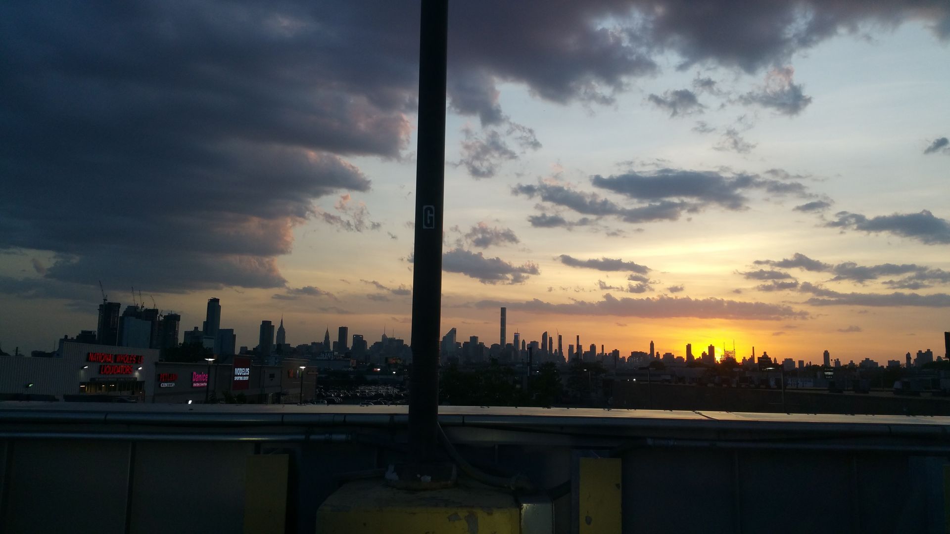 Sunset over a city skyline with cloudy sky and a pole in the foreground.