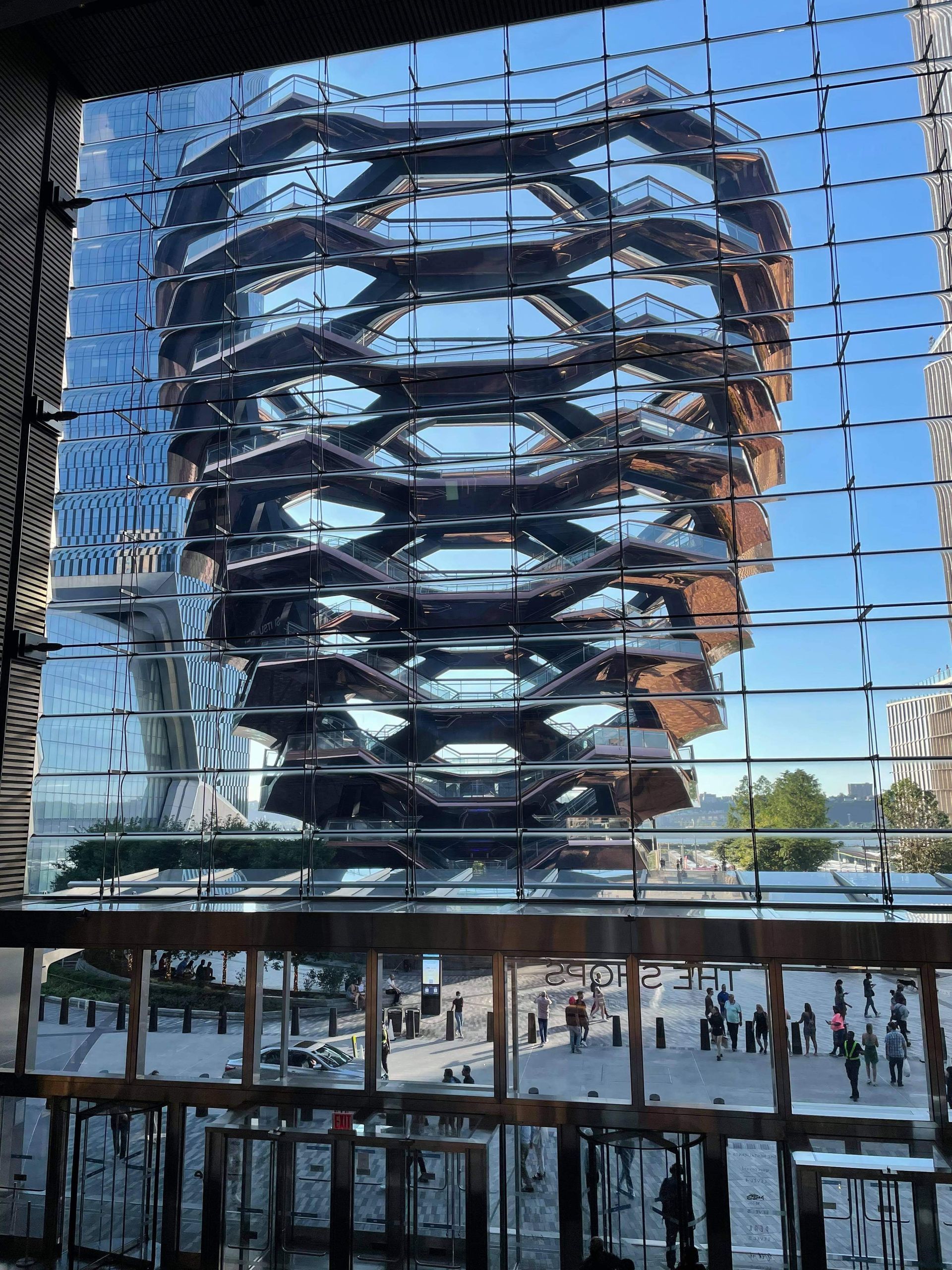 The Vessel, a honeycomb-shaped structure, reflected in a glass building's windows, in New York City. People walk in the plaza below.