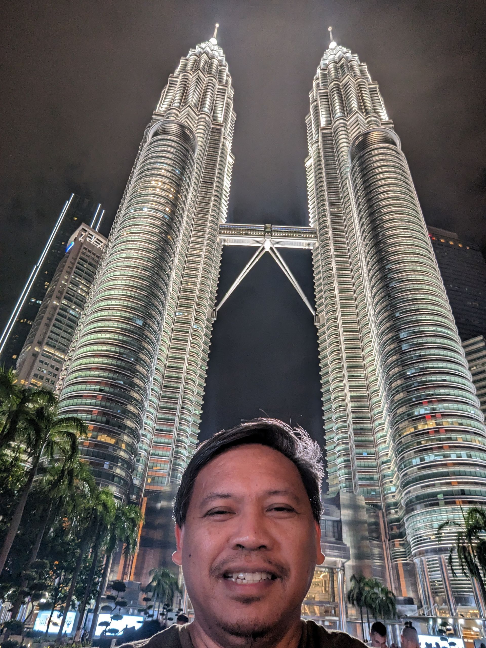 Man smiling in front of the illuminated Petronas Towers at night in Kuala Lumpur.