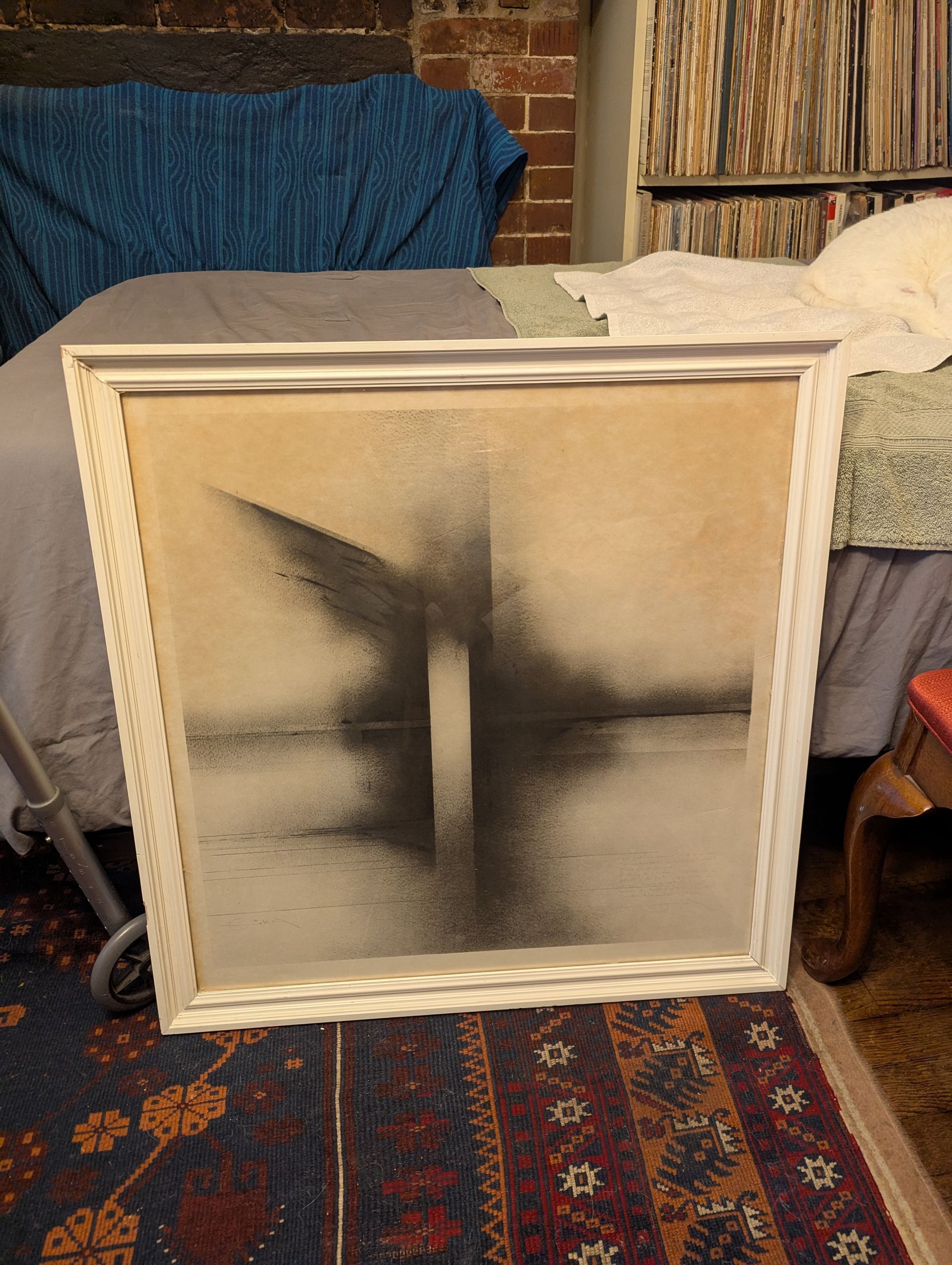 Abstract black and beige art in a white, ornate frame. Artwork stands on a patterned rug in front of a bed.