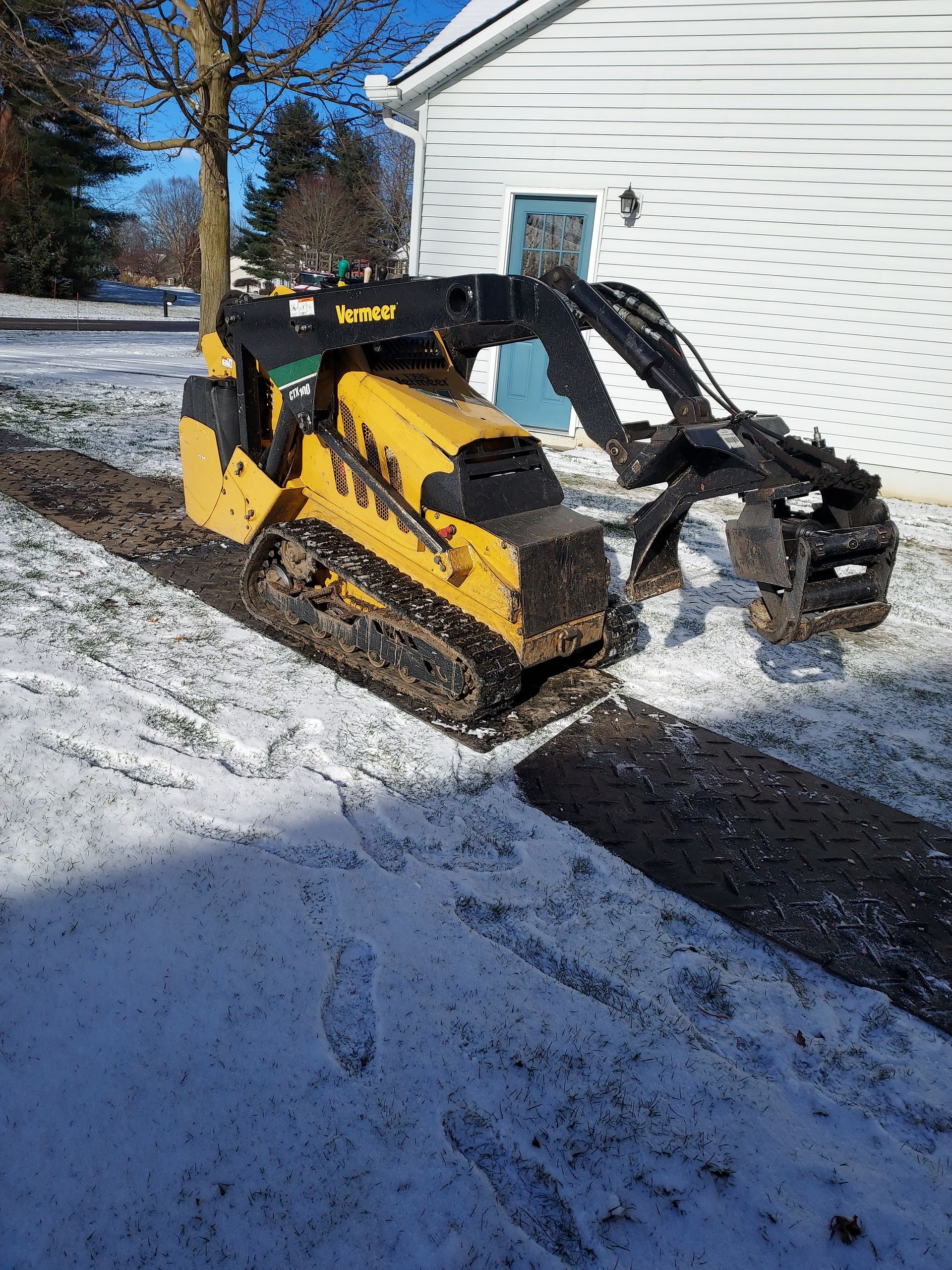 Yellow mini excavator digging a trench in snowy ground near a building.