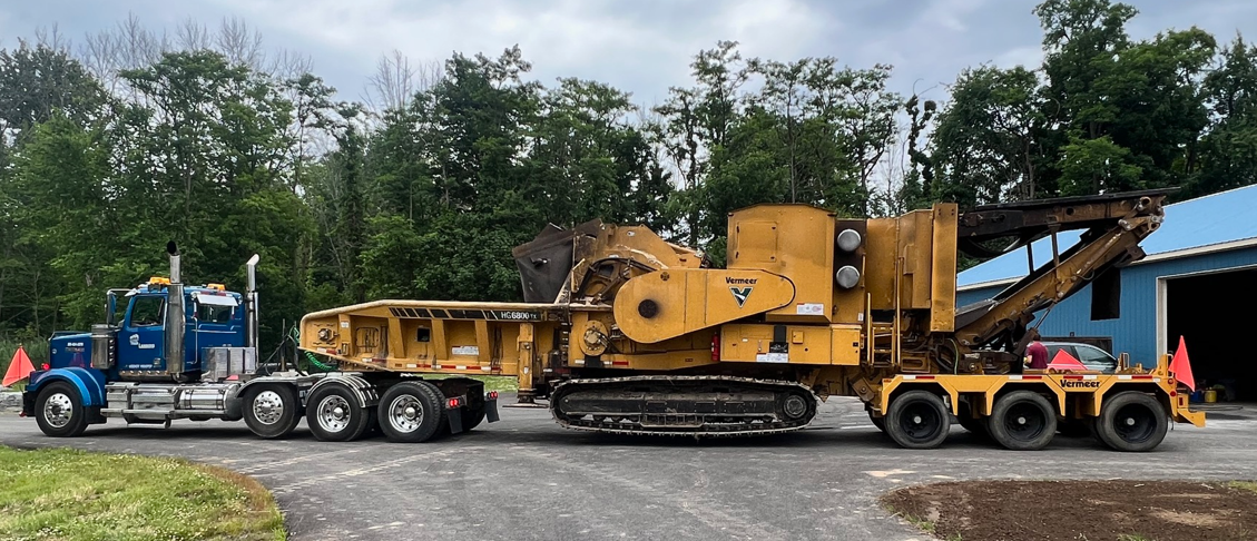 Blue semi-truck pulling yellow wood chipper on a road. Trees and a blue building are in the background.