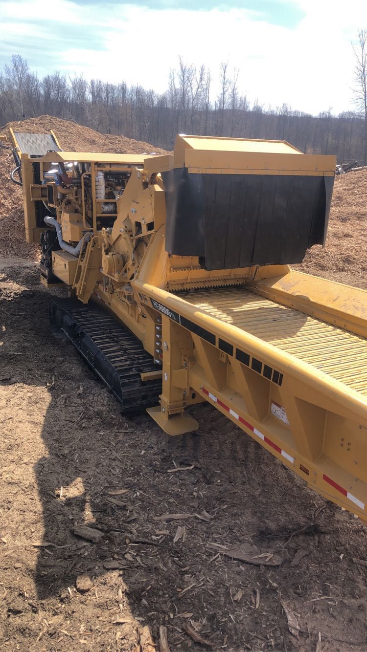 Yellow wood chipper on tracks in a wooded area, with conveyor belt extending outward.