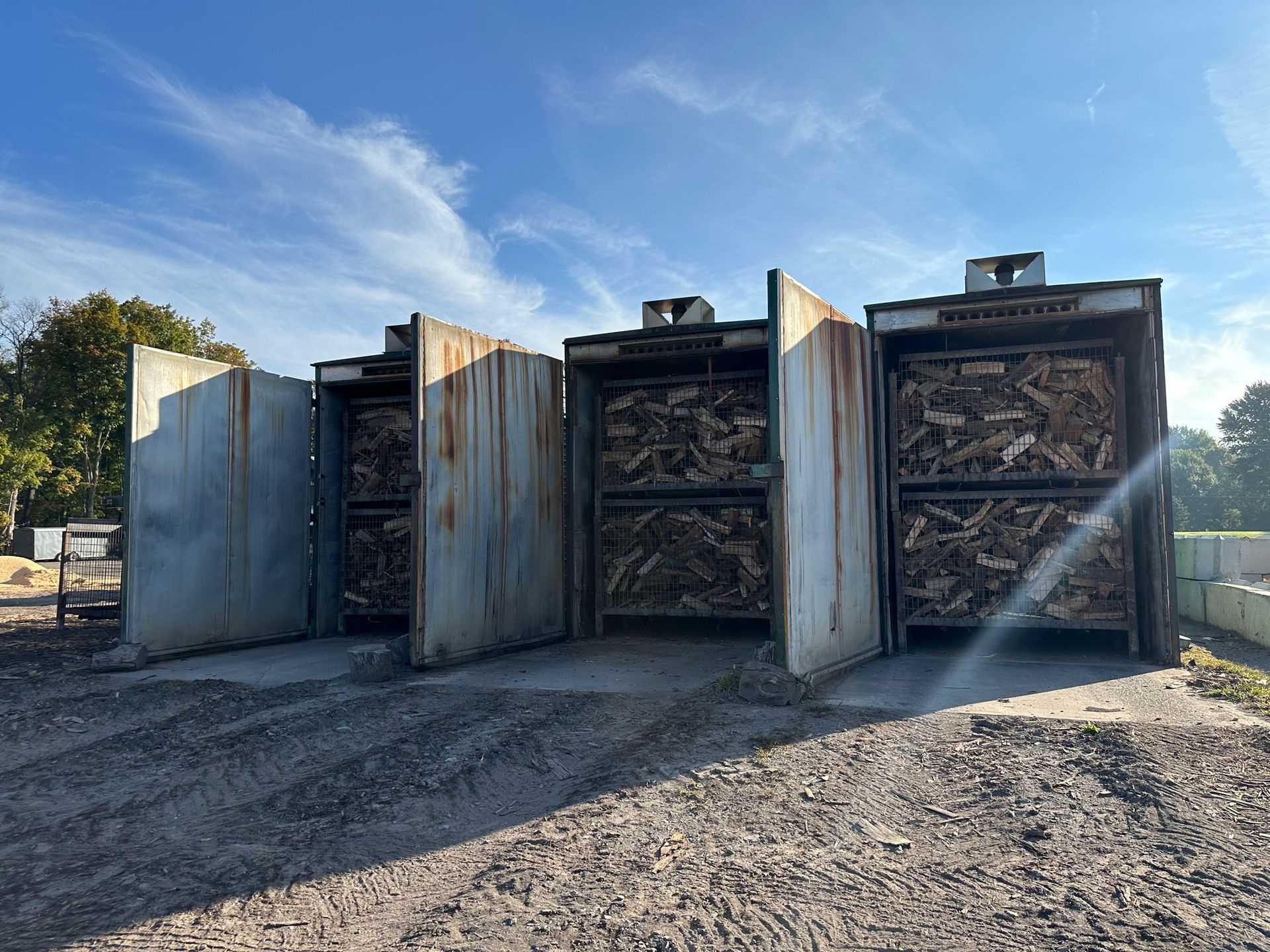 Three large metal drying kilns filled with wood chips, outdoors on a sunny day.