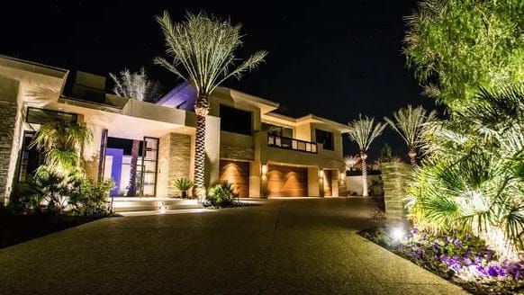 A large house is lit up at night with palm trees in front of it.