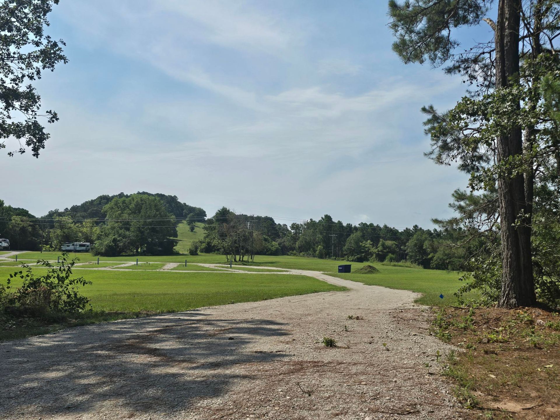 Gravel path through a sunny green park with trees and a pond under a blue sky