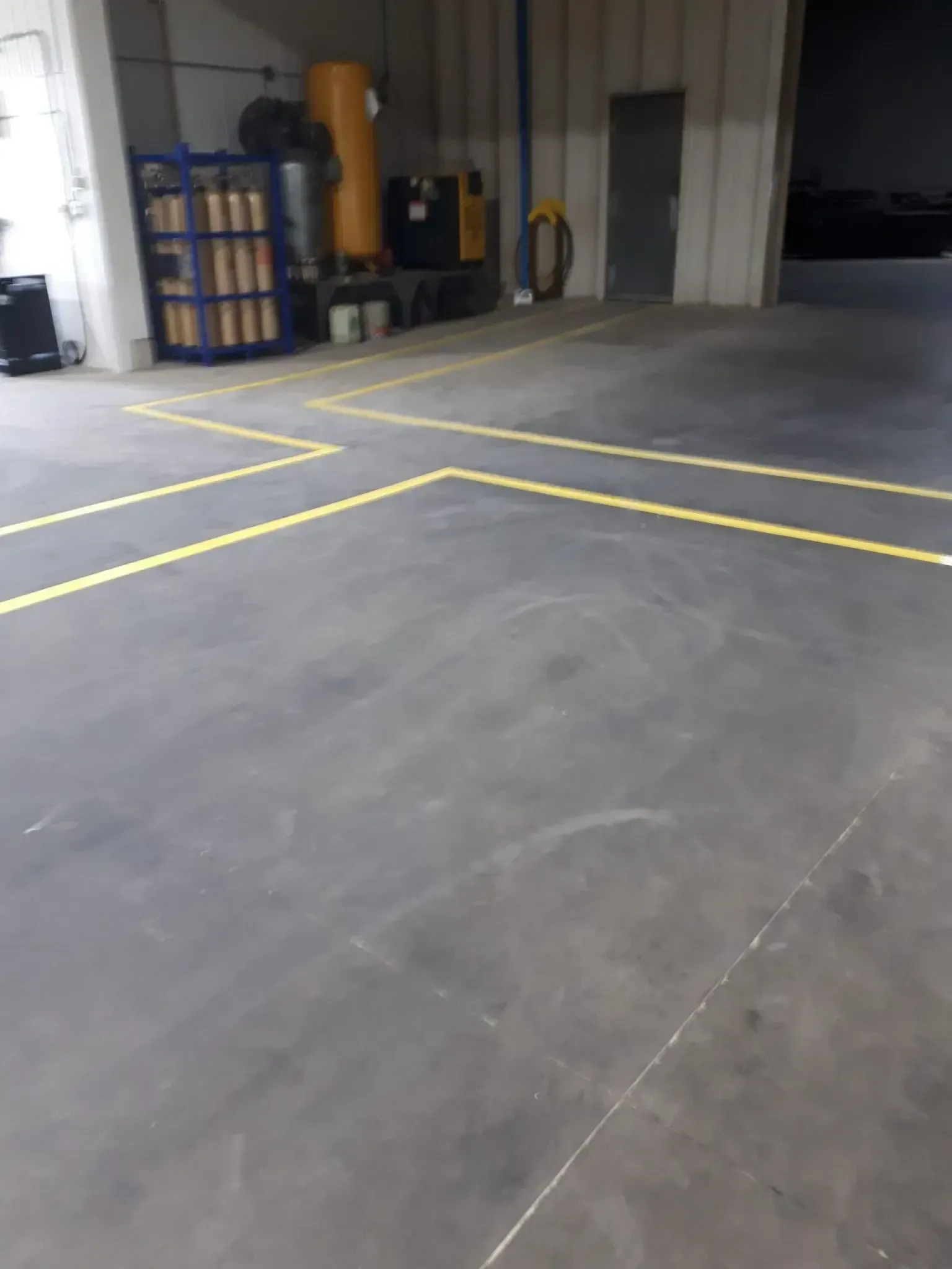 Empty warehouse floor with yellow lines. Tools and equipment line a wall; a door is in the background.