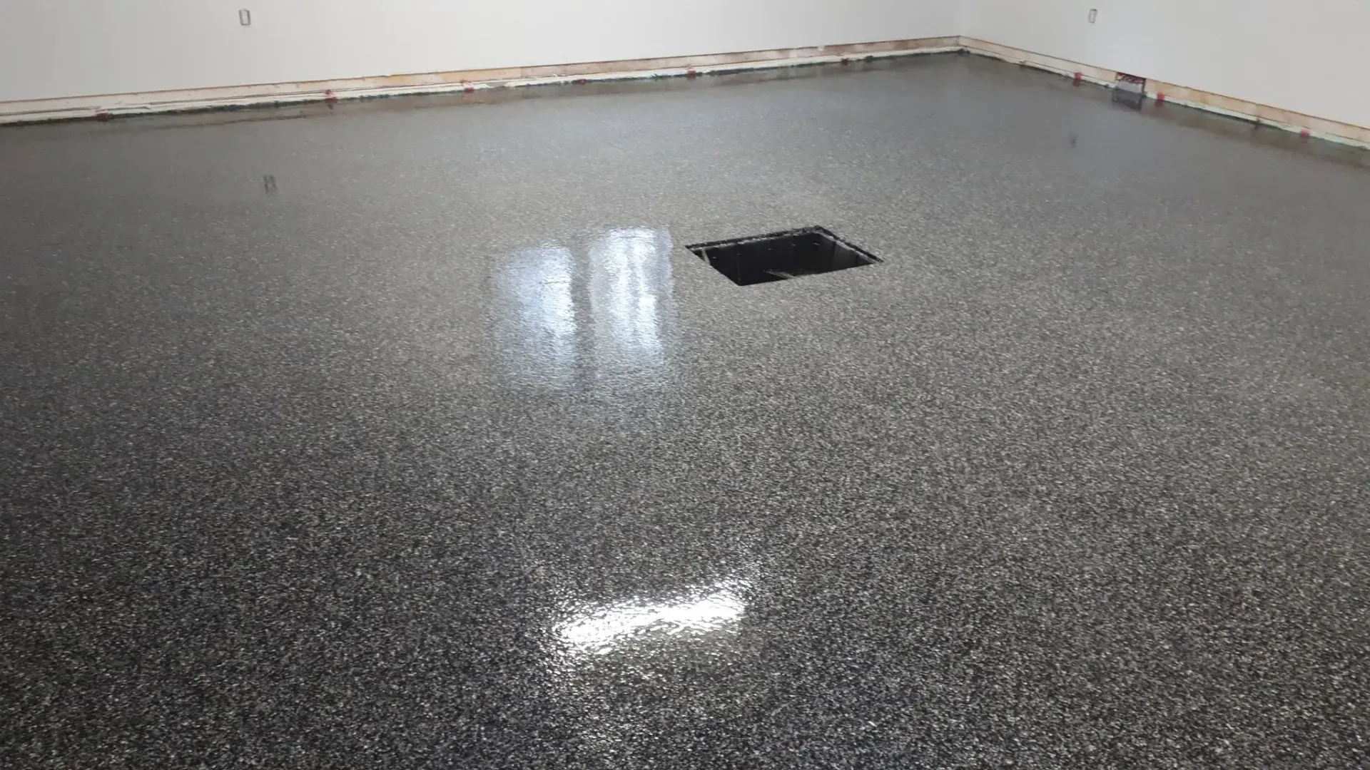 A glossy, grey epoxy floor with black and white flecks, featuring a dark square drain in the center of a room.