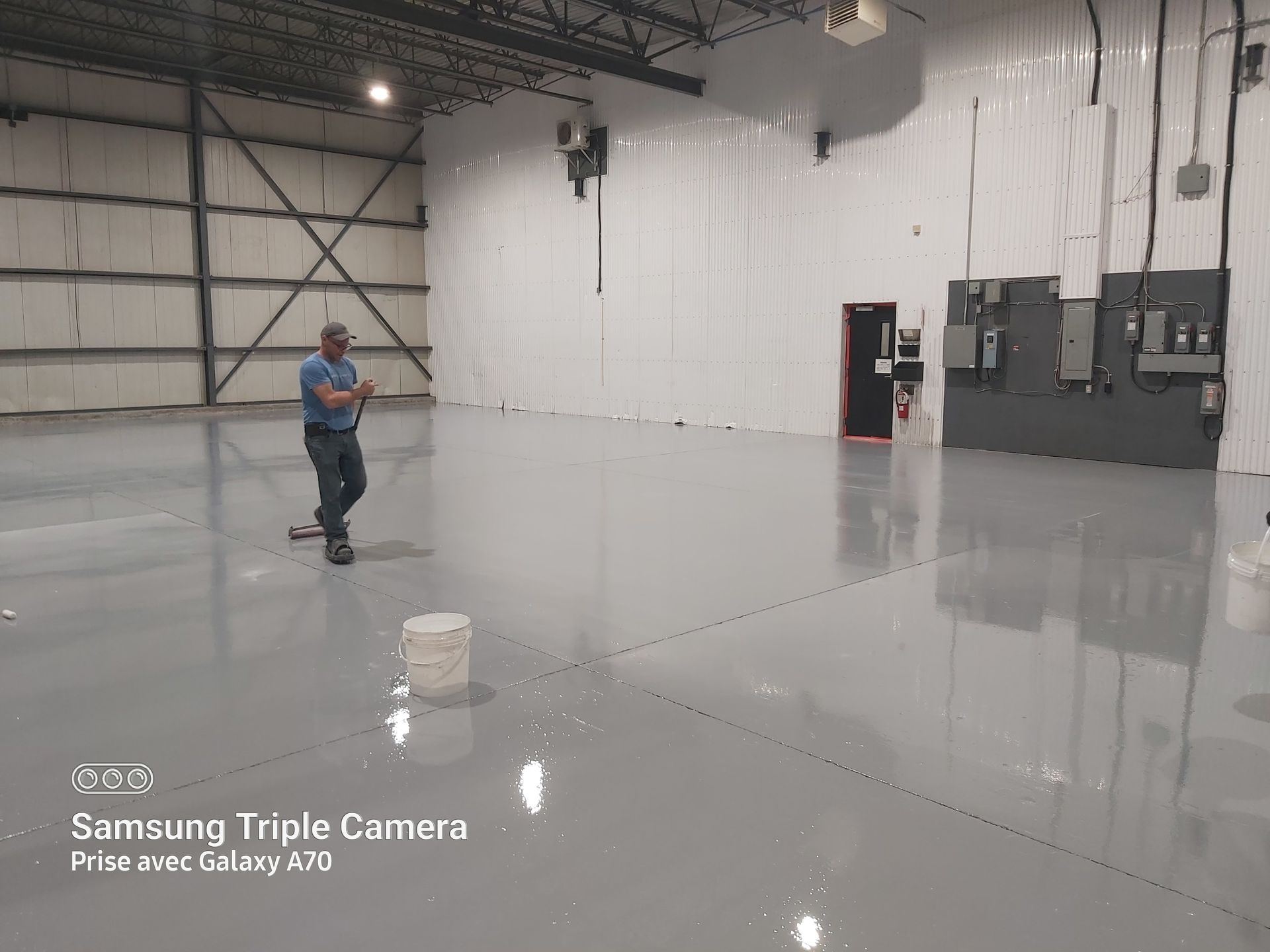 Man walking in a large, empty gray-floored warehouse, holding a tool; white walls and metallic beams overhead.