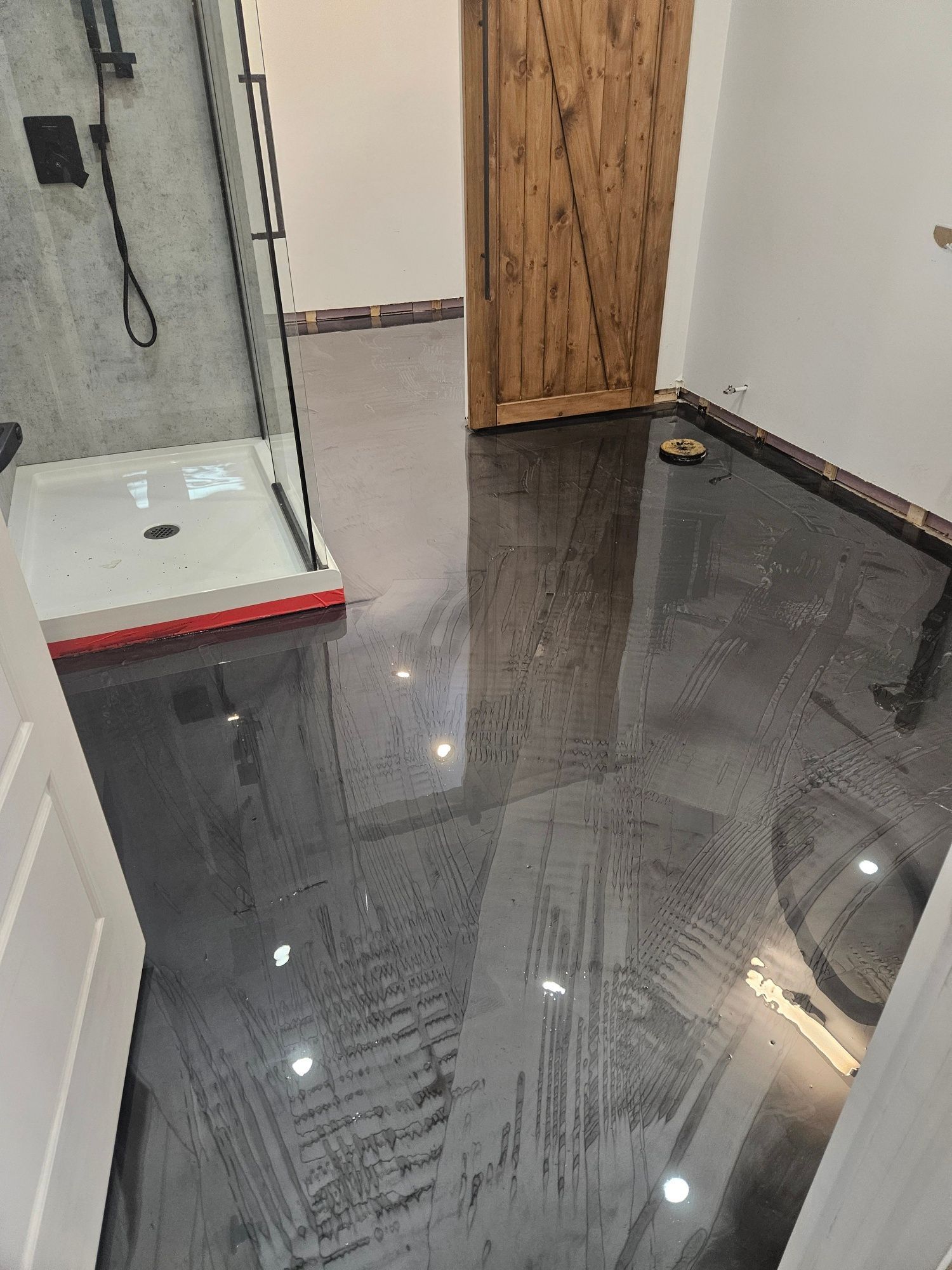 Bathroom with shiny black epoxy floor, glass shower, wooden door, and white walls.
