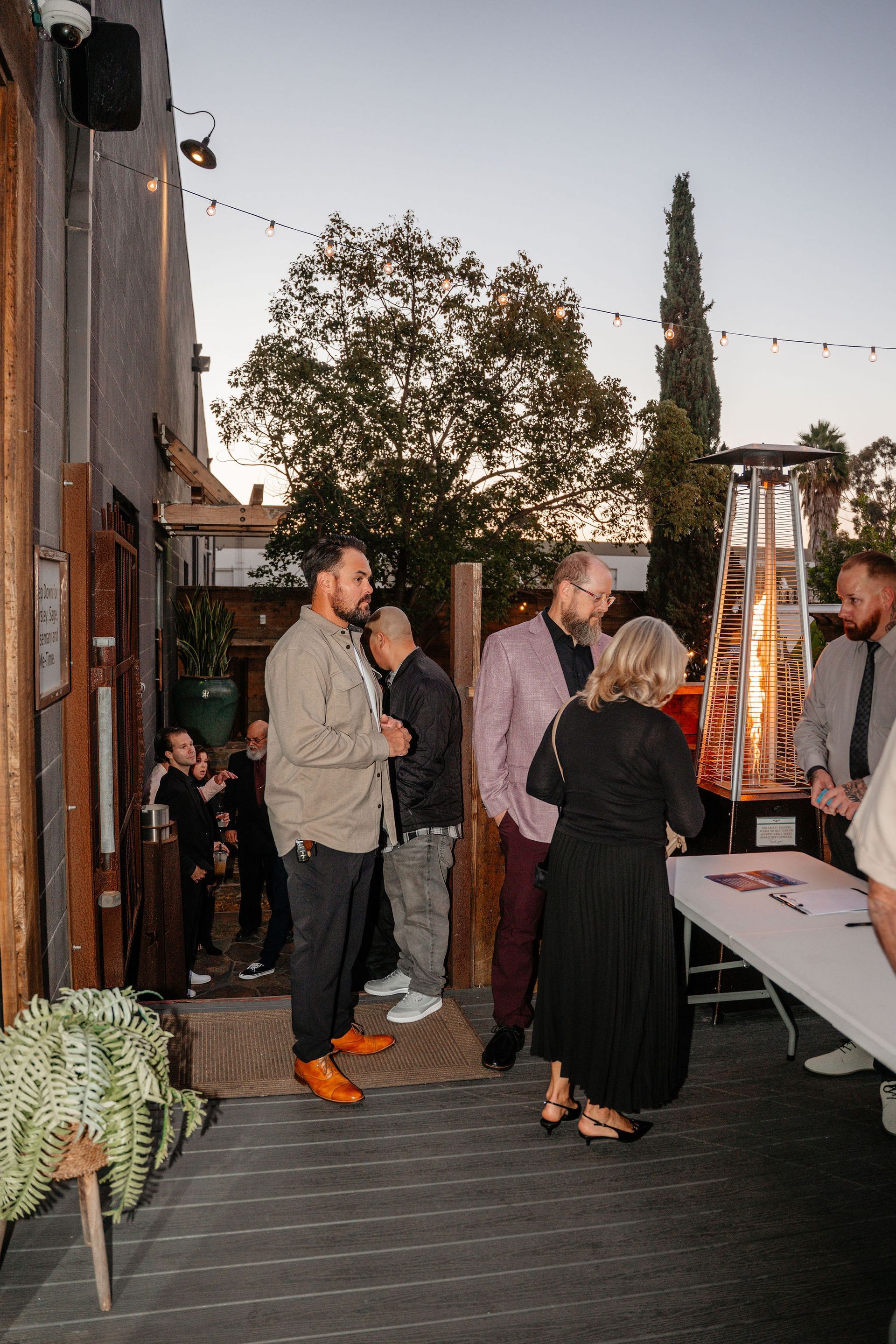 People gather outside a building, near a registration table and a lit heater. Evening setting.