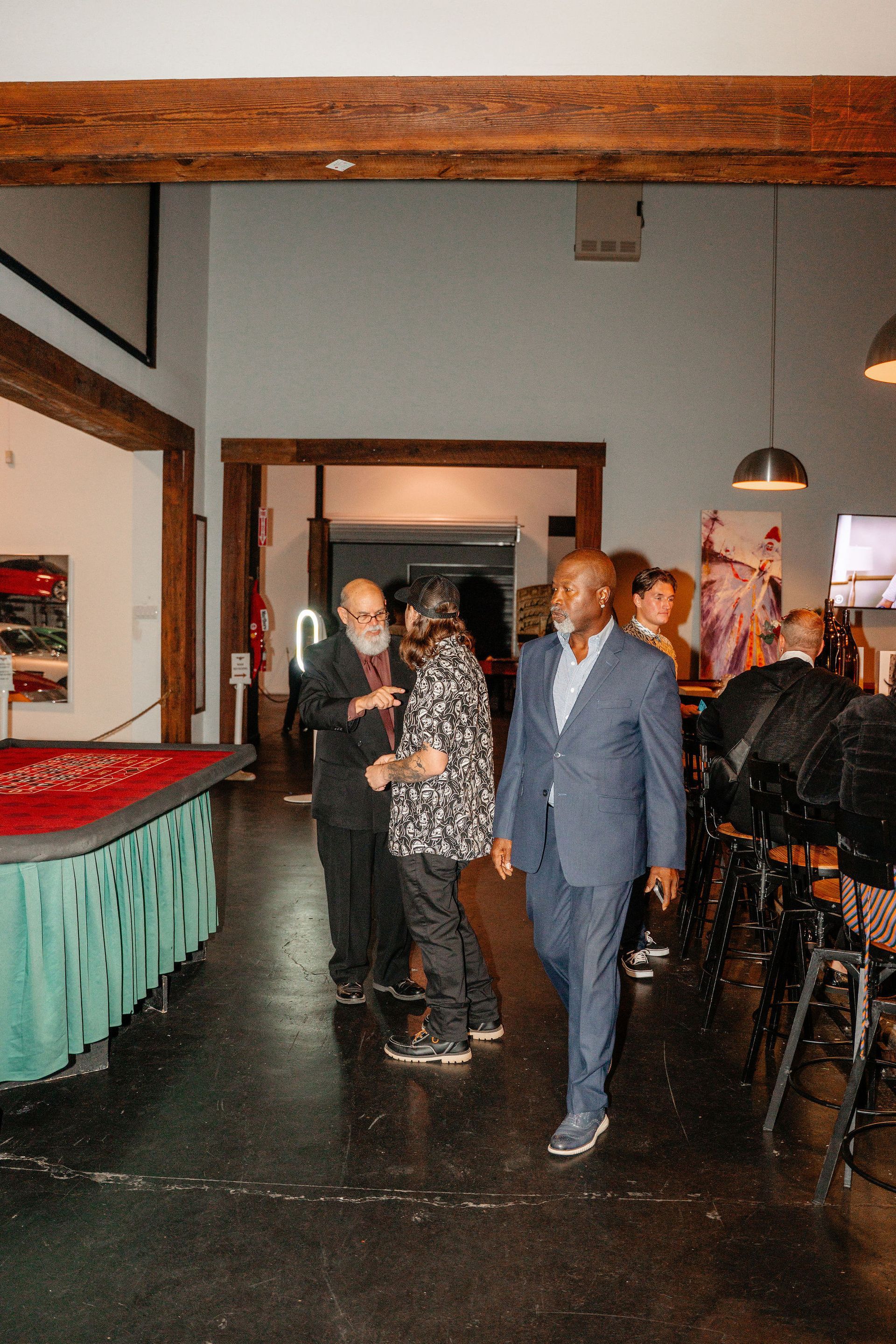 Man in blue suit walks through a bar. Two men converse in the background near a roulette table.