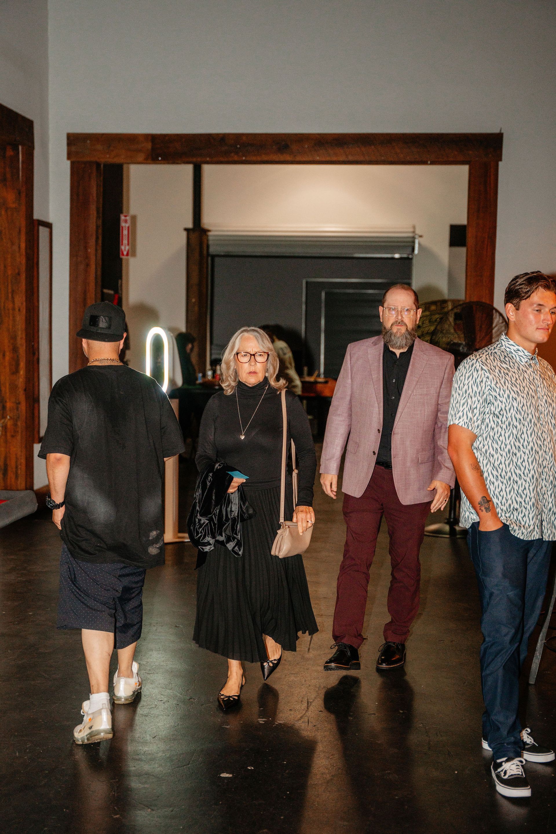 People in a building. Woman in black dress, man in pink blazer, other guests.