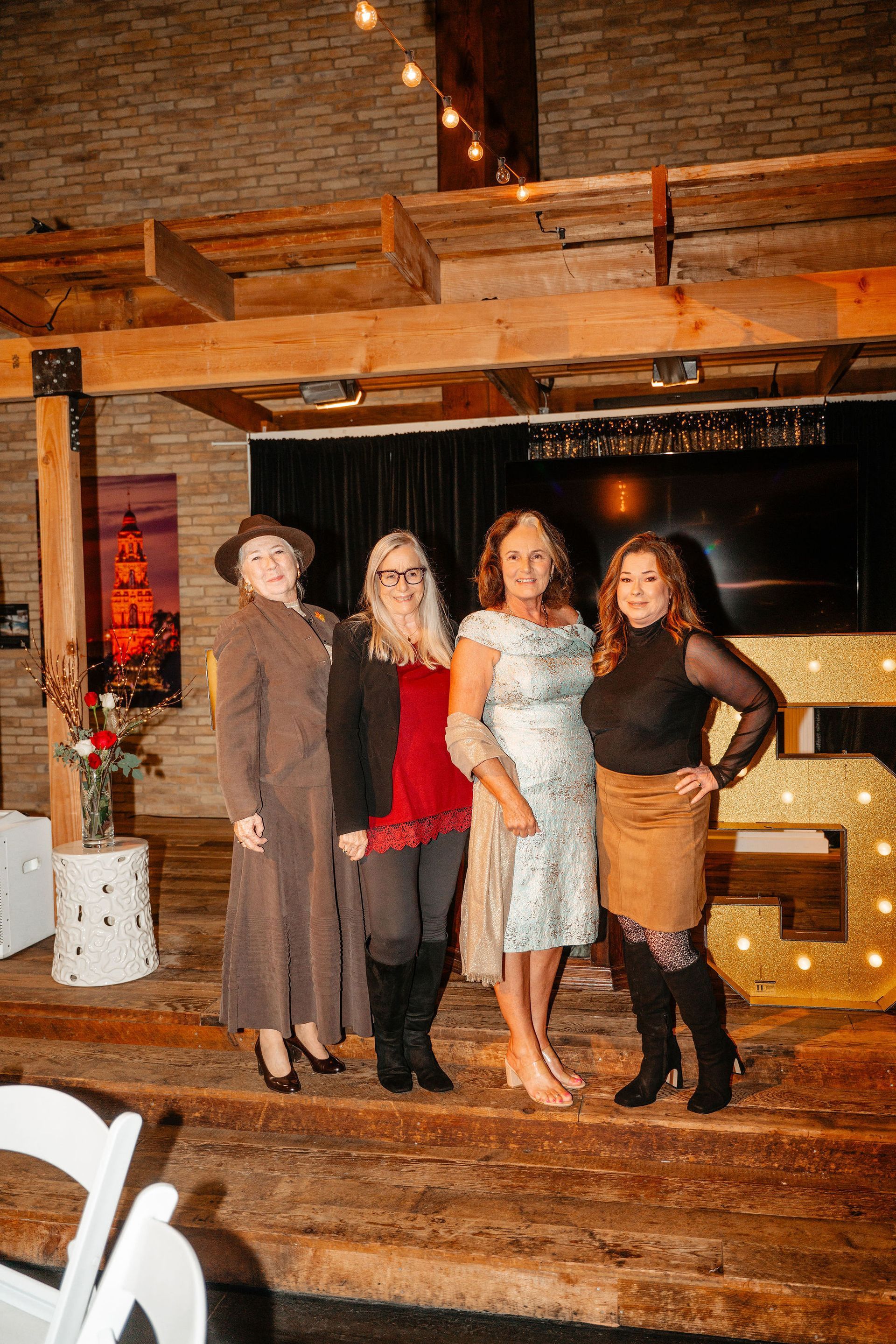 Four women smiling, posing at an event. Gold “50” sign. Rustic wood and brick setting, party lights.