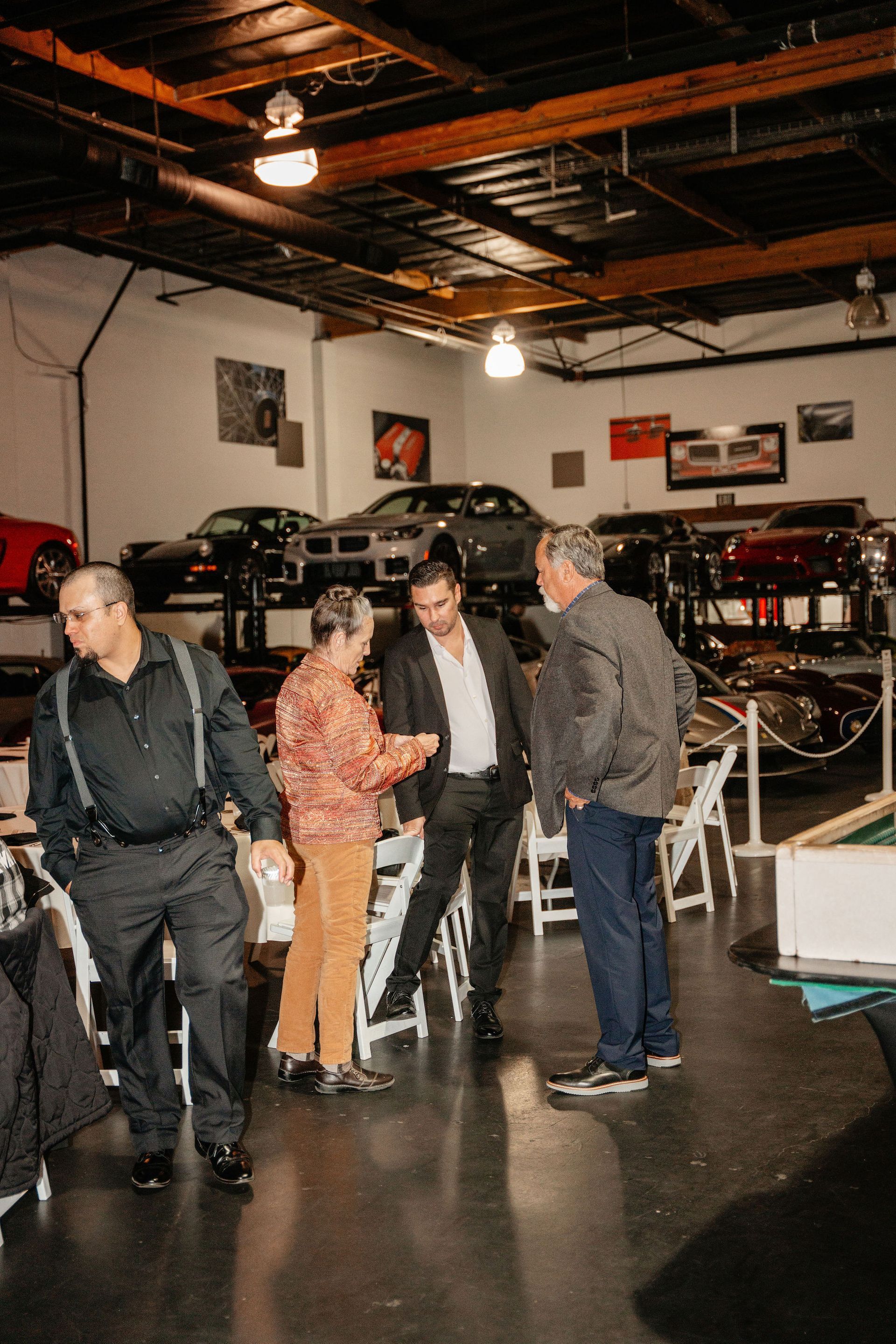 Four men conversing inside a garage with classic cars on display. Two are looking at something in a man's hand.