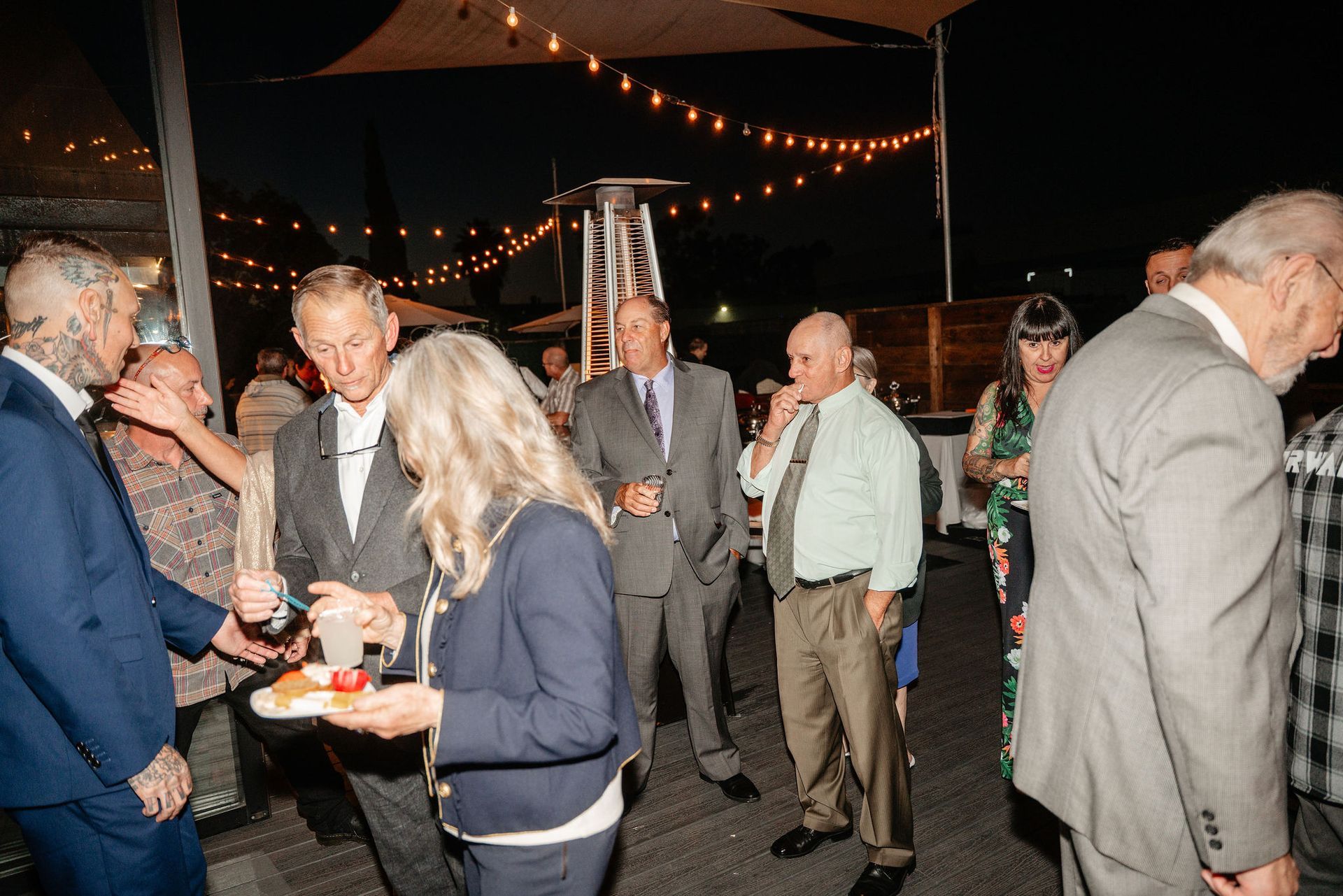 People socialize at an outdoor evening gathering under string lights; some are eating, wearing suits, and talking.