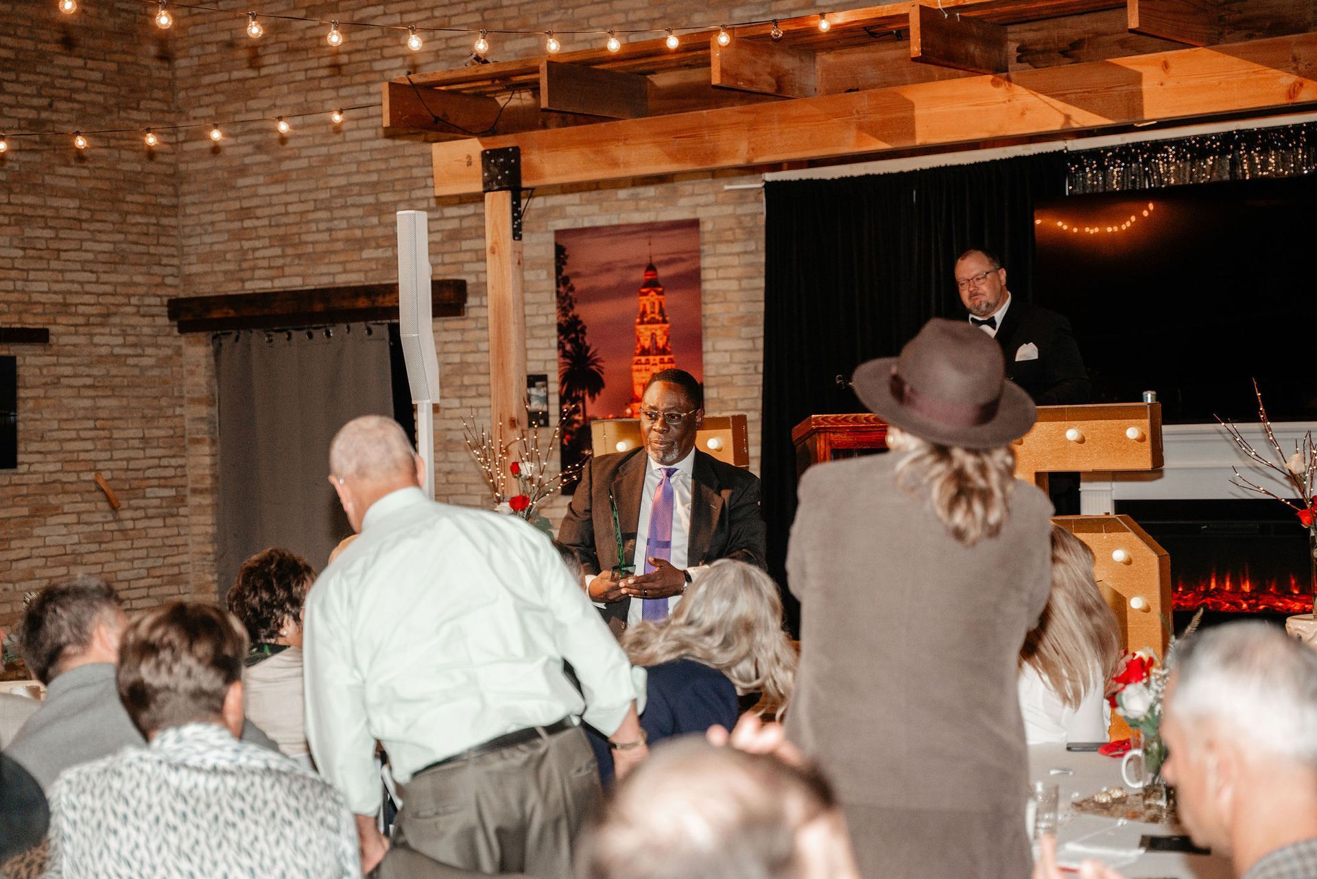 People at a formal event; a man speaks at a podium. Brick wall and stage in background.