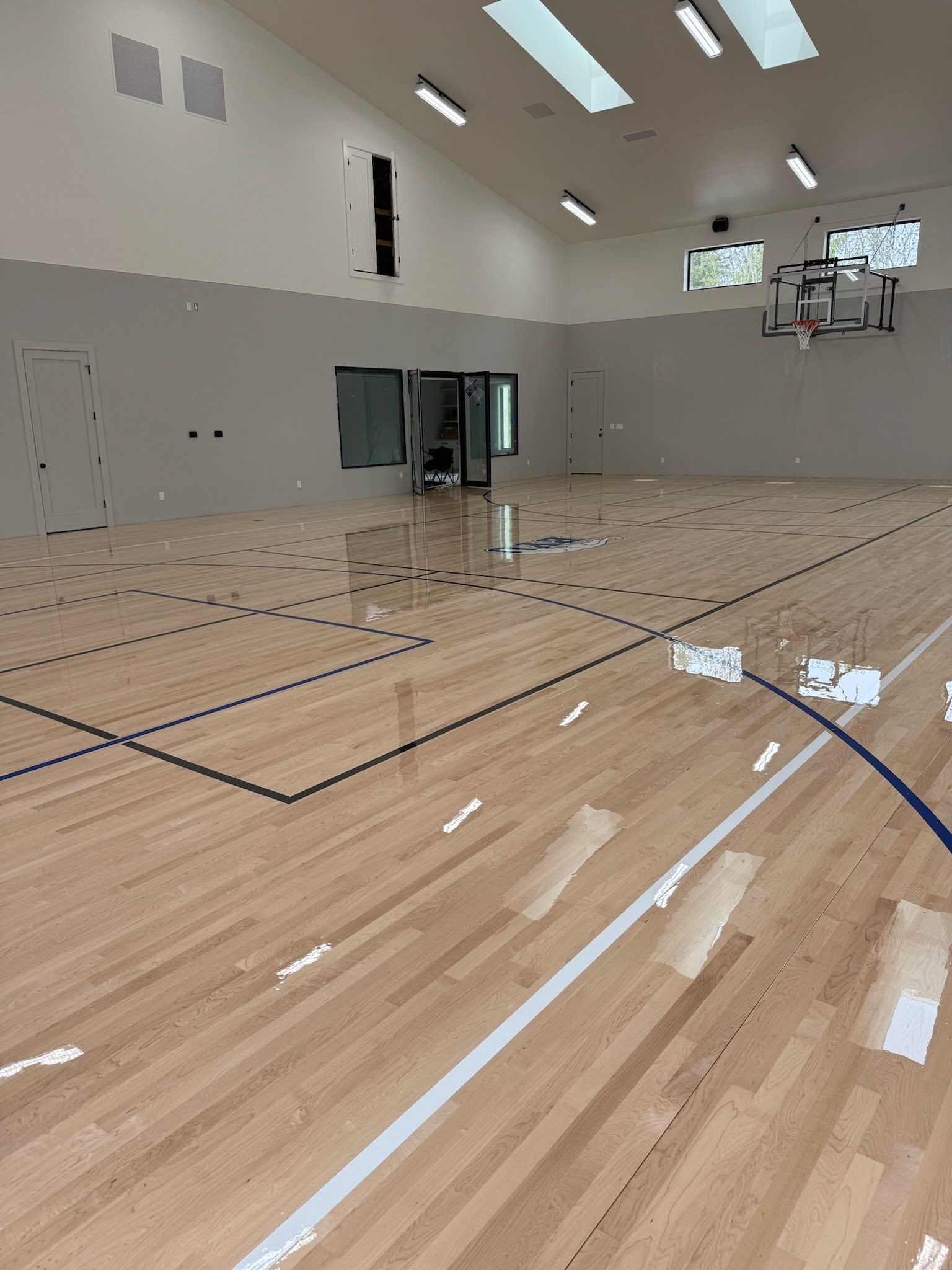 Indoor residential basketball court or gymnasium with a hoop, vaulted ceiling, and multiple windows/skylights.
