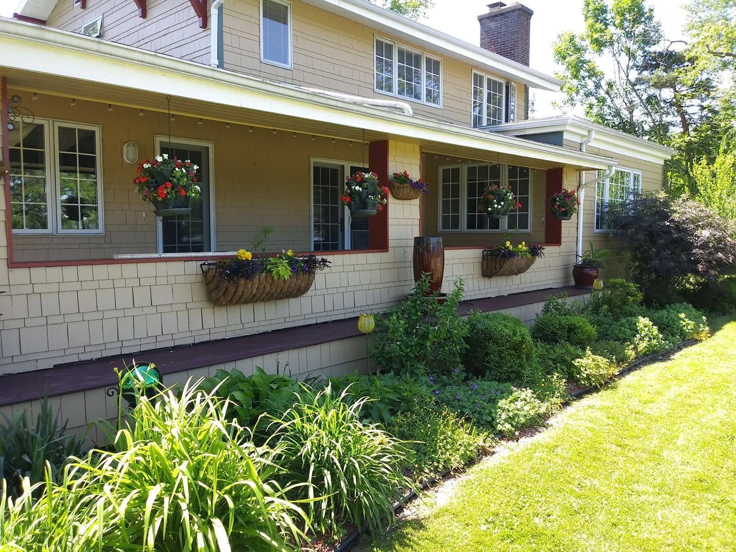 Front porch of a beige house with flower boxes, hanging plants, and a lush green garden in sunlight