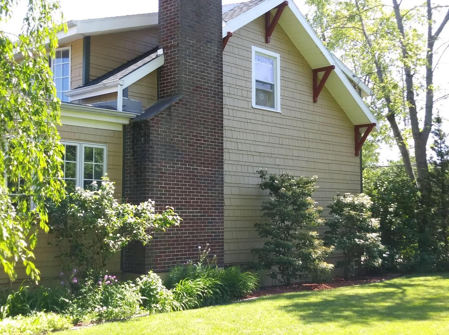 Suburban house with tan siding, brick chimney, and trees in a sunny yard