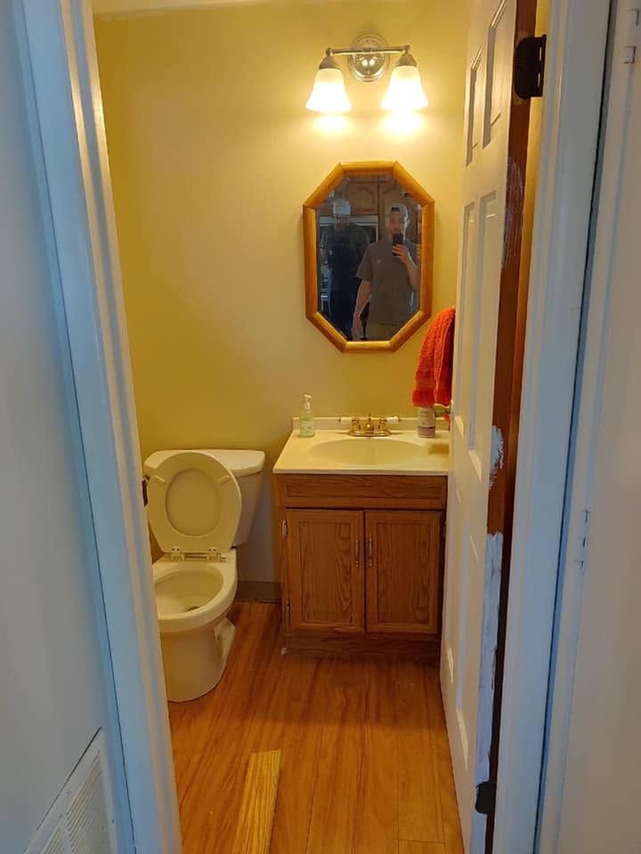 Small bathroom with a toilet, sink, mirror, and warm yellow walls and wood-look floor.