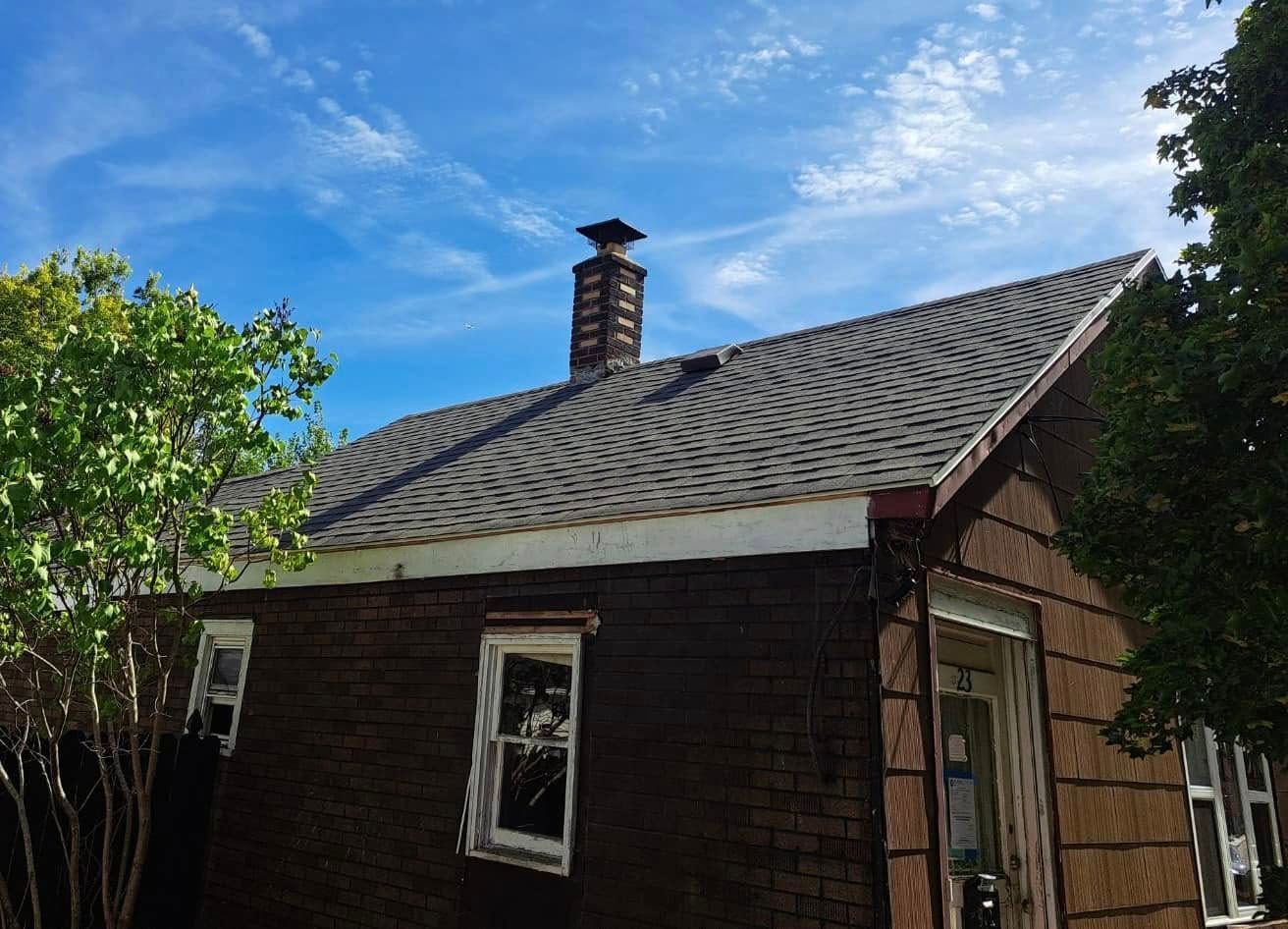 Small dark wooden house with a shingled roof and chimney under a blue sky, framed by green trees