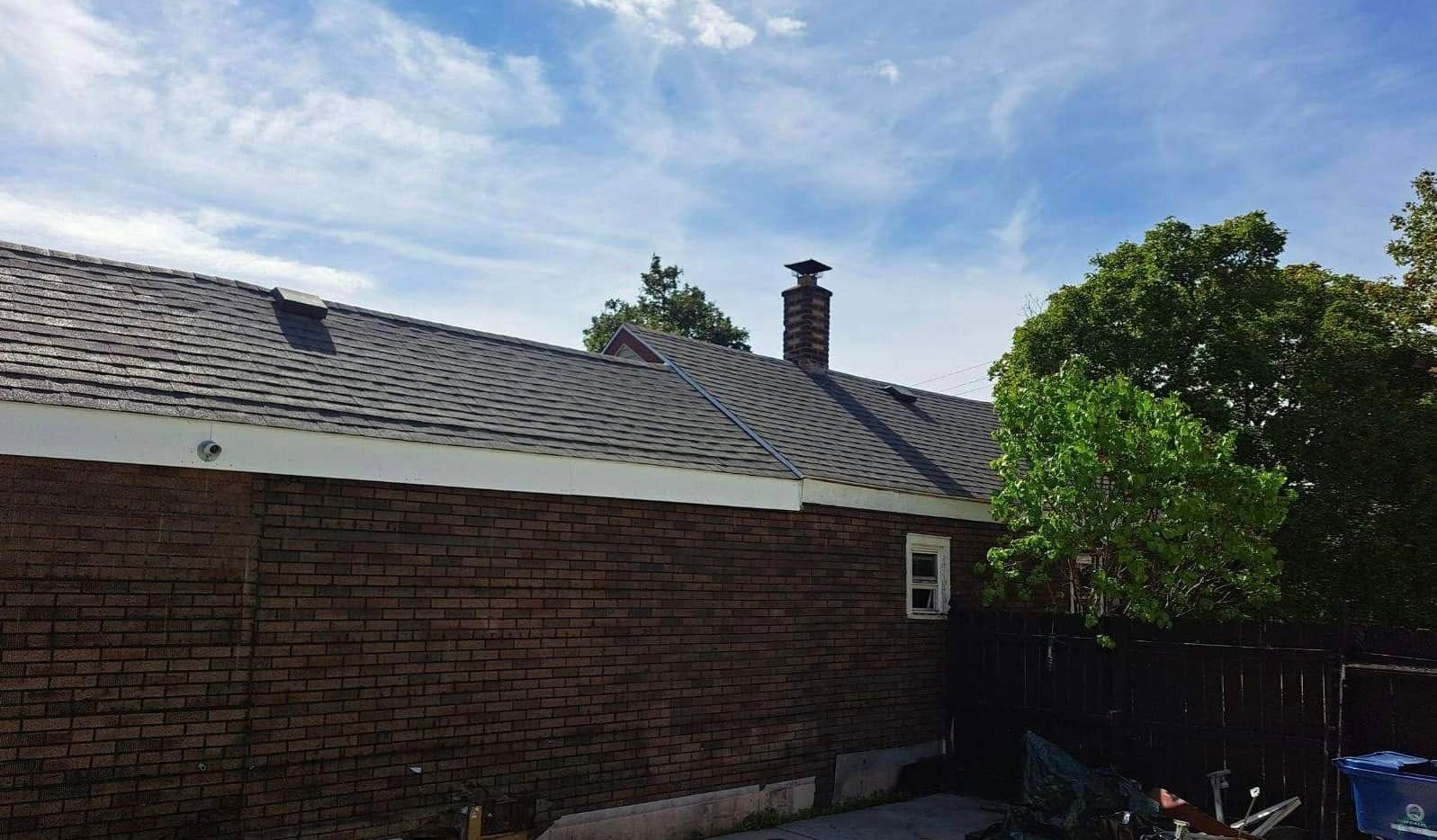 Backyard view of a brick house with a gray shingle roof, chimney, and trees under a blue sky