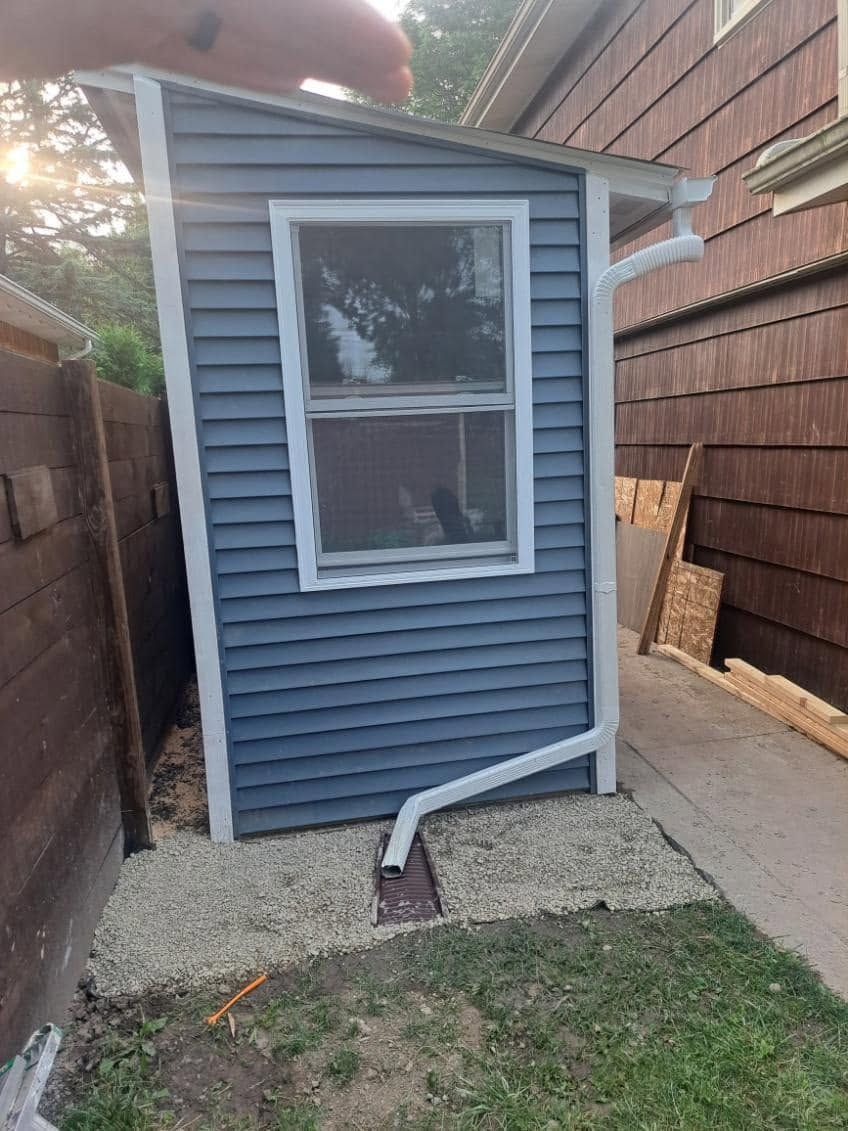 Small blue shed with white trim beside a brick wall and gravel path, with a hose in front.