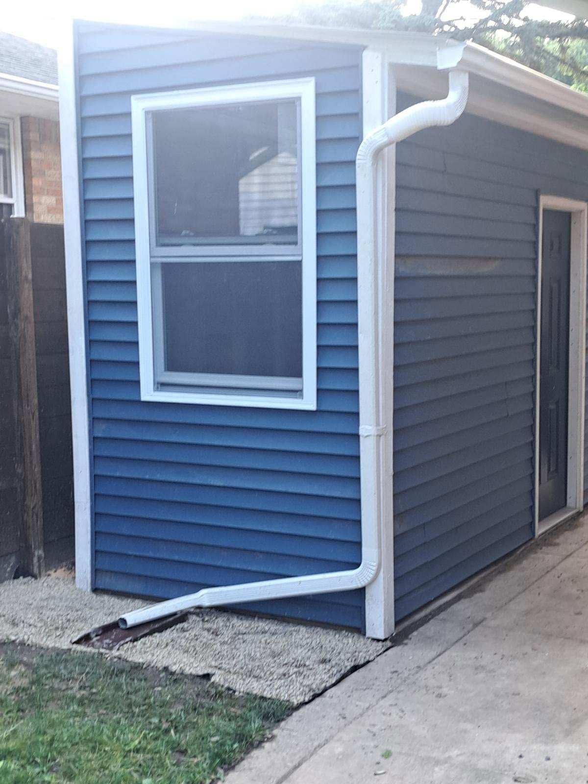 Small blue house exterior with white trim, window, gutter, and side door beside a concrete walkway.