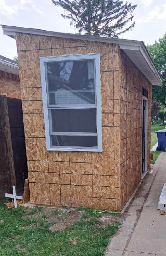Small unfinished shed with plywood siding and a white-framed window beside a driveway.