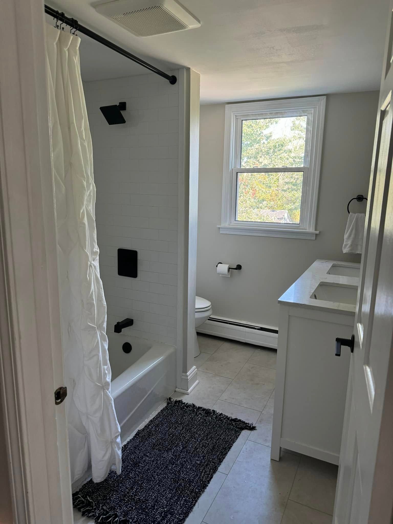 Small bathroom with a shower, vanity, window, and dark rug on the floor.