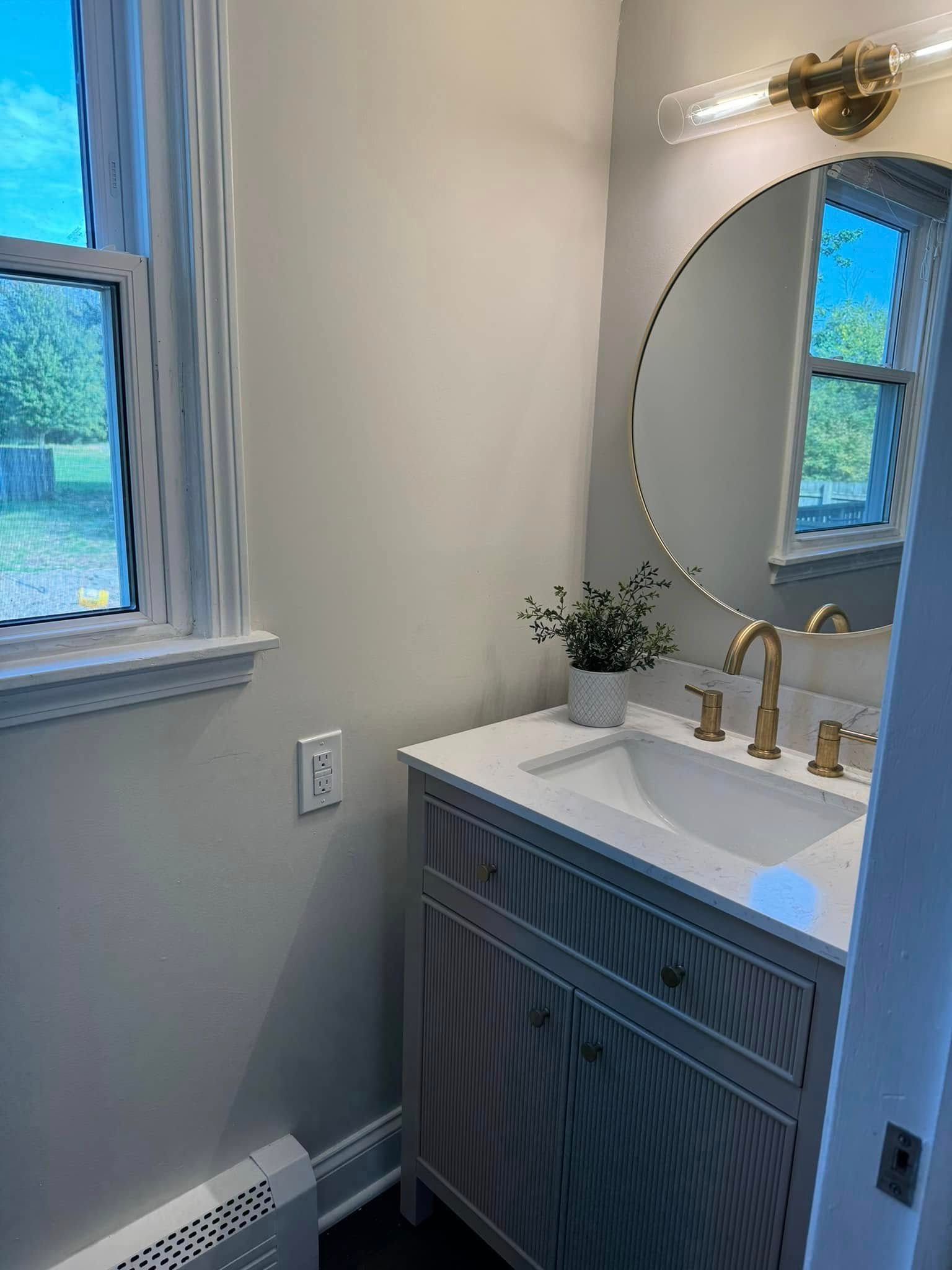 Small bathroom vanity with round mirror, brass light, white sink, and window beside a blue cabinet.