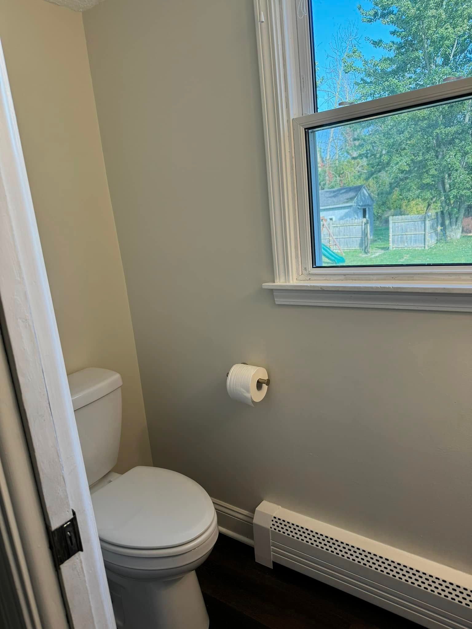 Small bathroom with toilet, toilet paper roll, radiator, and window overlooking a yard.