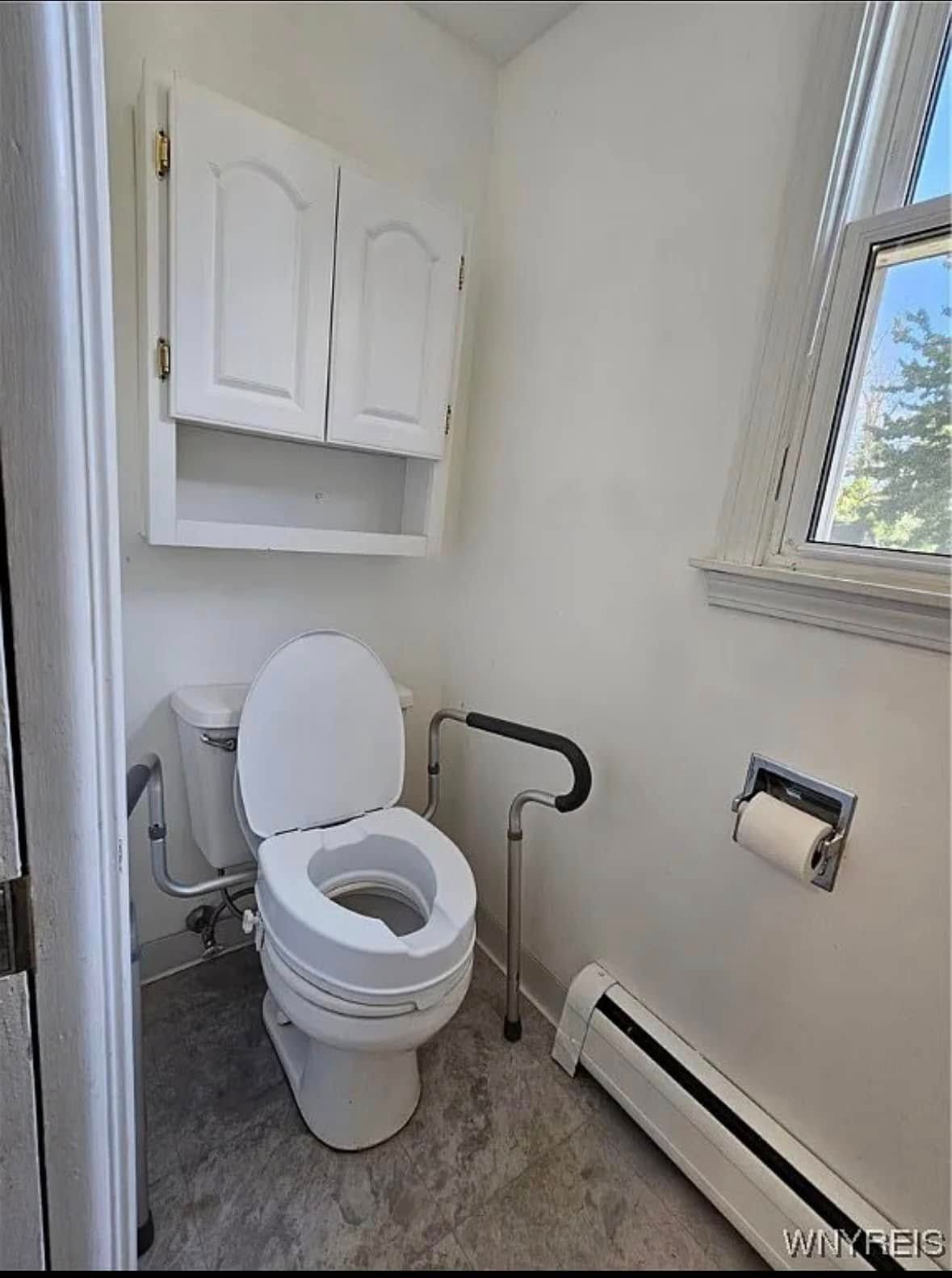 Small white bathroom with toilet, wall cabinet, window, and radiator.