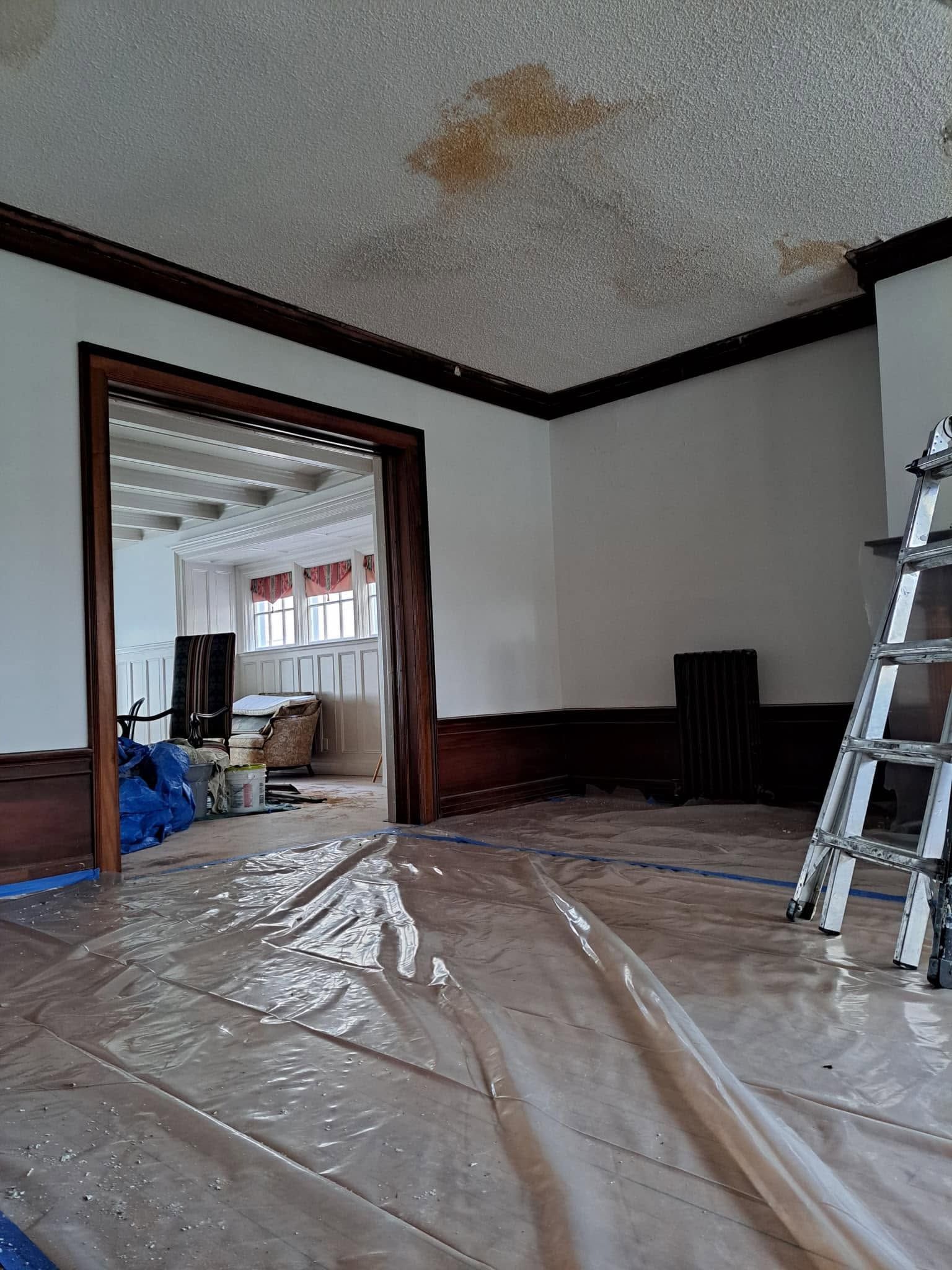 Empty room under renovation with plastic-covered floor, ladder, doorway to another room, and ceiling stain
