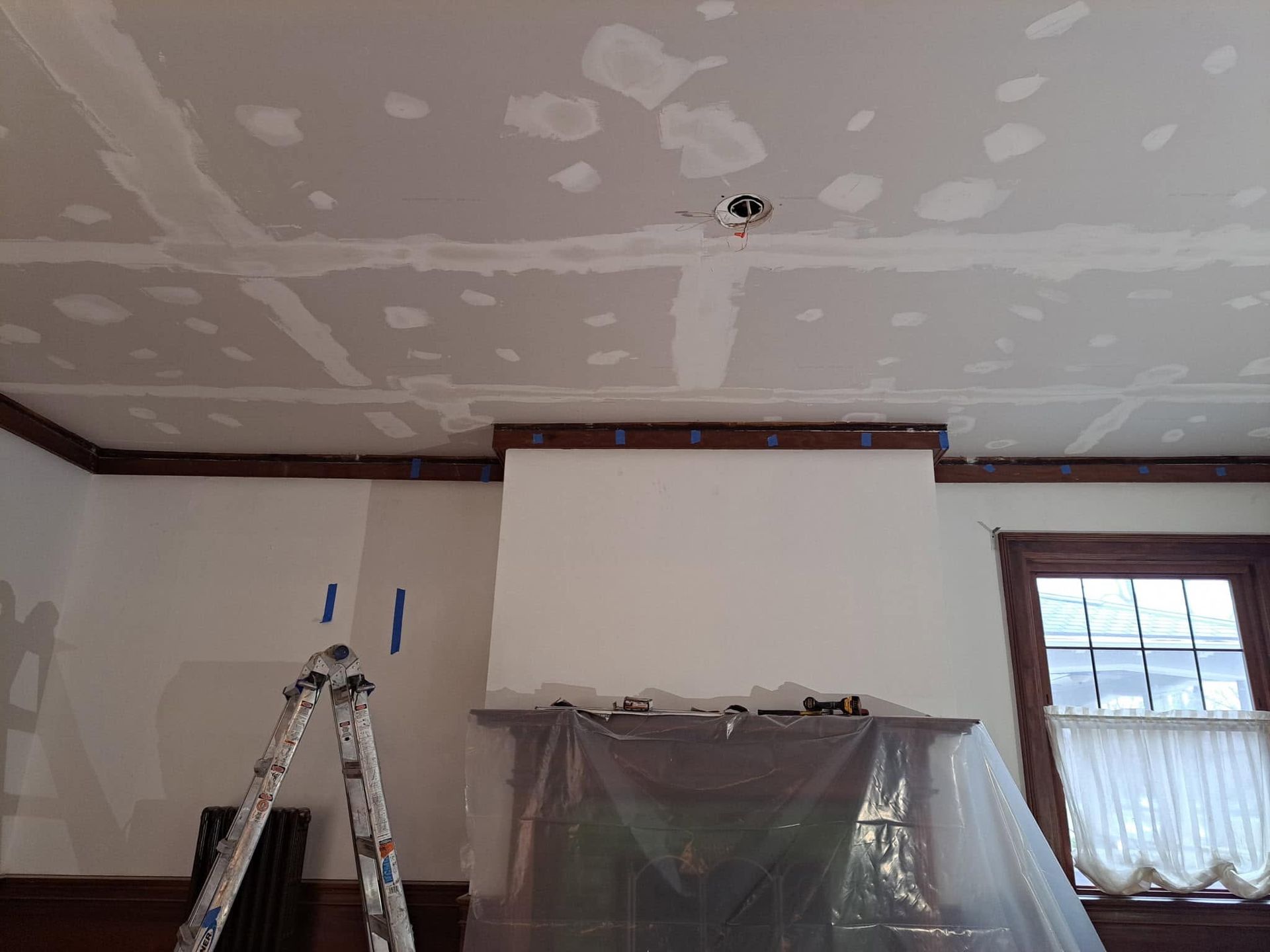 Room under renovation with patched ceiling, ladder, and plastic-covered furniture near a window.