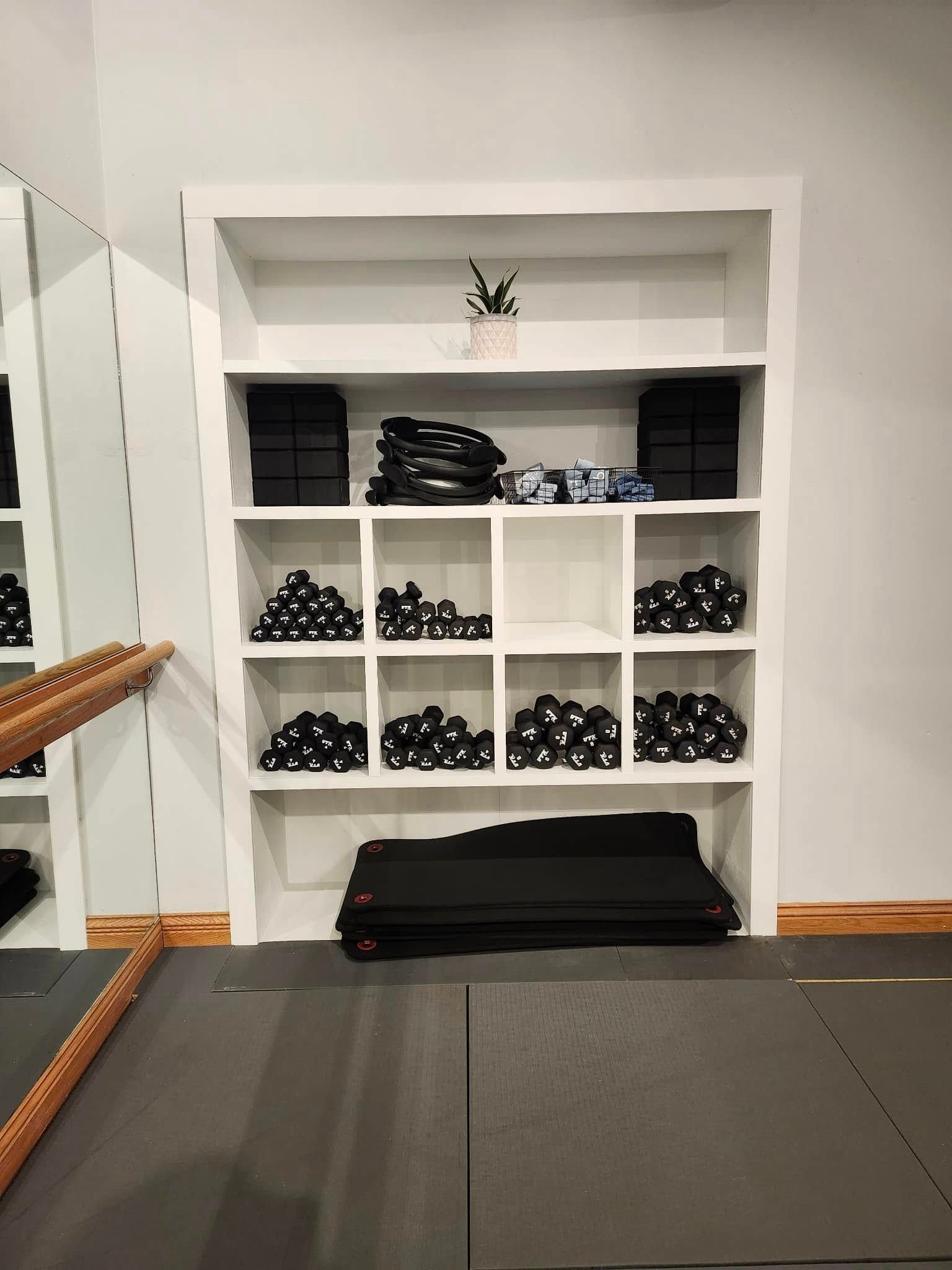 Gym studio shelves with stacked black workout props and folded mats beside a mirror.