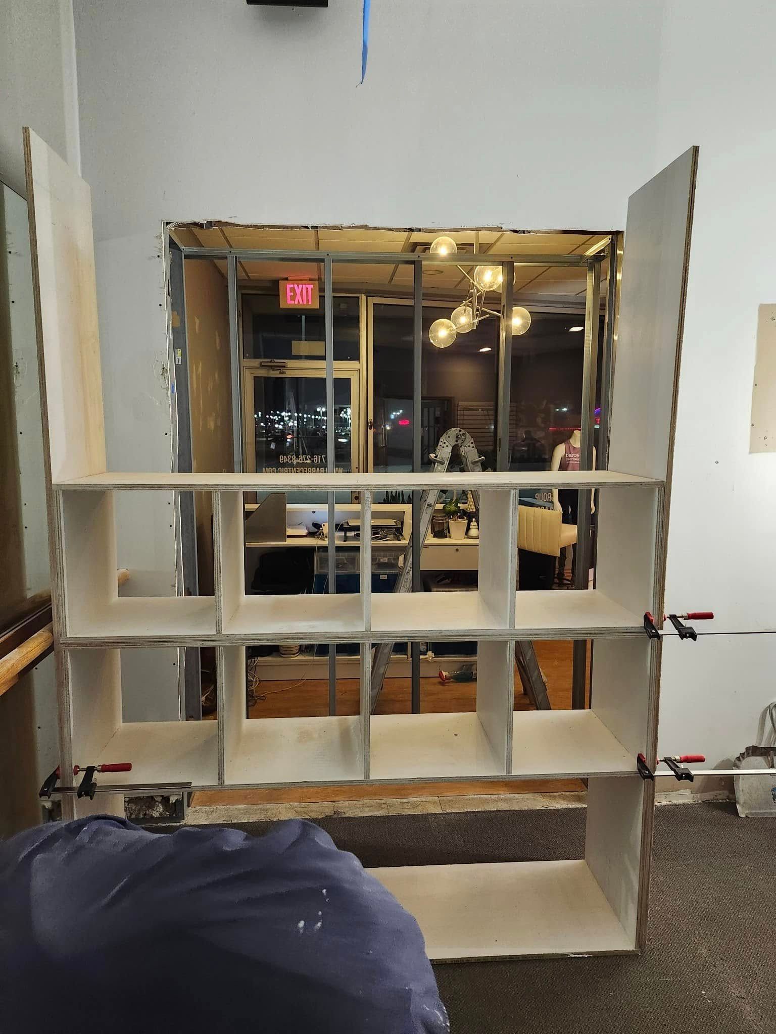 A white display shelf by a storefront window in a carpeted indoor space, with a dark bag in the foreground.
