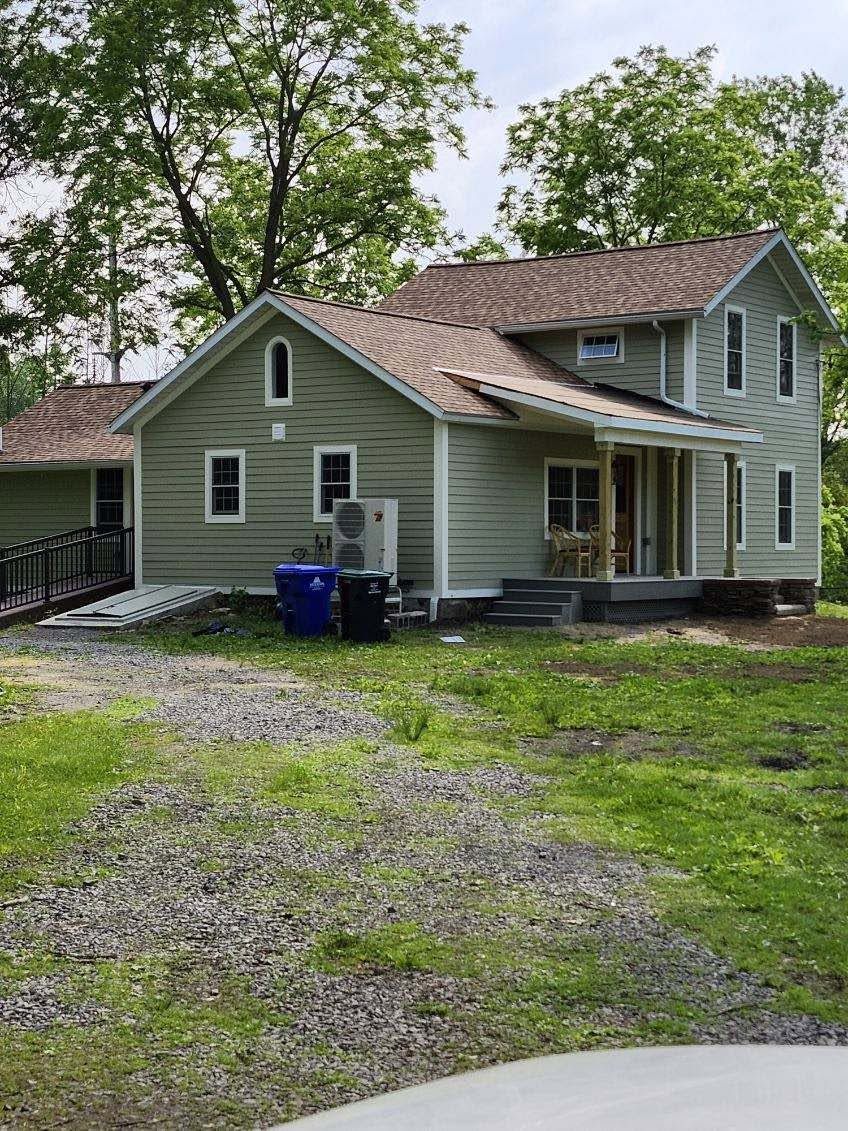 Green two-story house with a front porch, gravel driveway, and trees in the yard