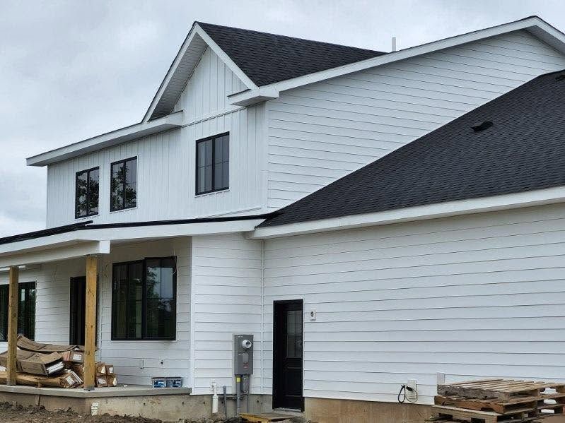 White two-story house under construction with black roof and unfinished porch framing