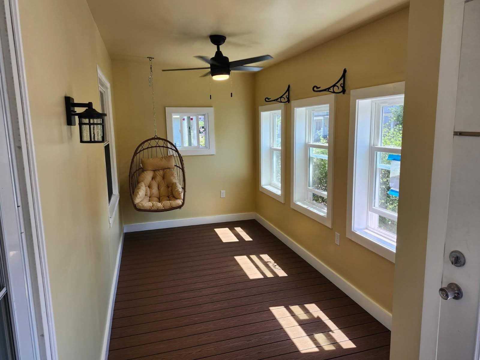Narrow beige sunroom with windows, ceiling fan, hanging chair, and brown carpet runner.